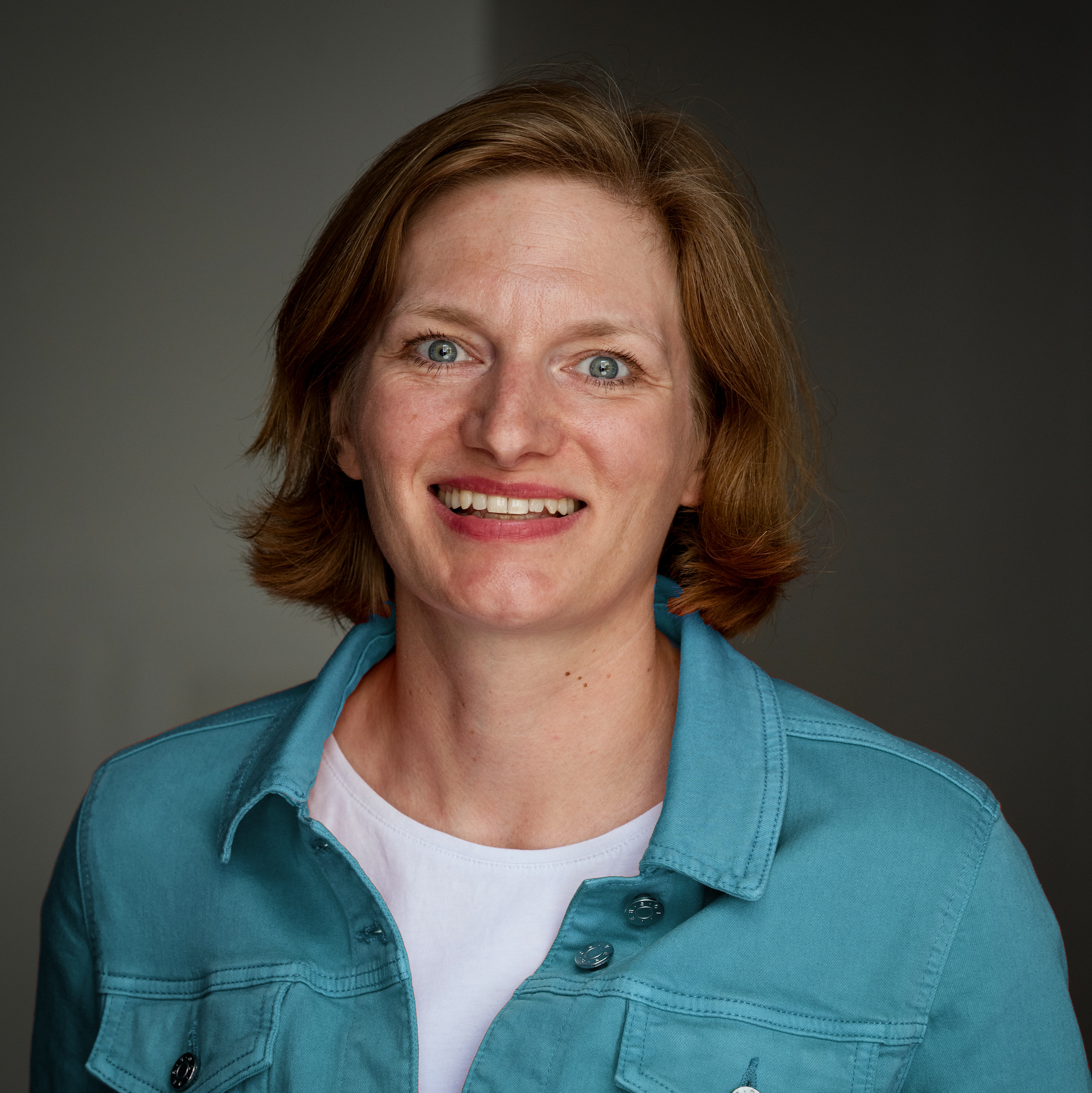 Ein Porträtfoto einer lächelnden Frau mit roten Haaren, blauen Augen, in einer blauen Jacke und weißem T-Shirt gegen einen grauen Hintergrund.