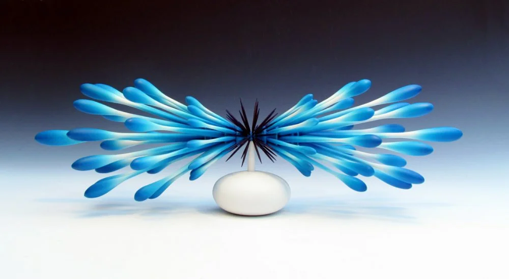 A decorative sculpture with a white oval base and a black spiky element in the center, surrounded by blue elongated petal-like structures radiating outward.