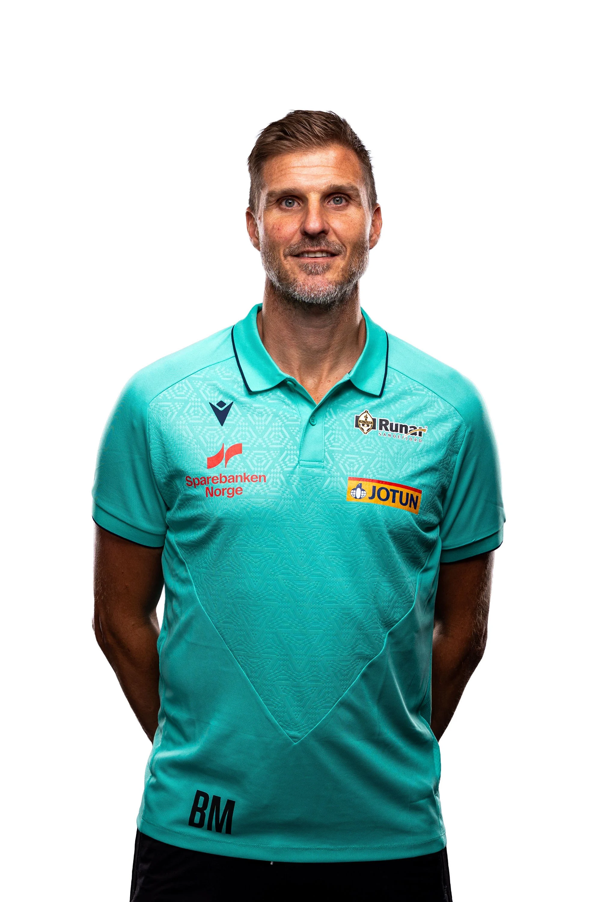En mann med brunt kort hår og skjegg, iført en lyseblå sportsleder med flere sponsorlogoer, står mot en hvit bakgrunn.