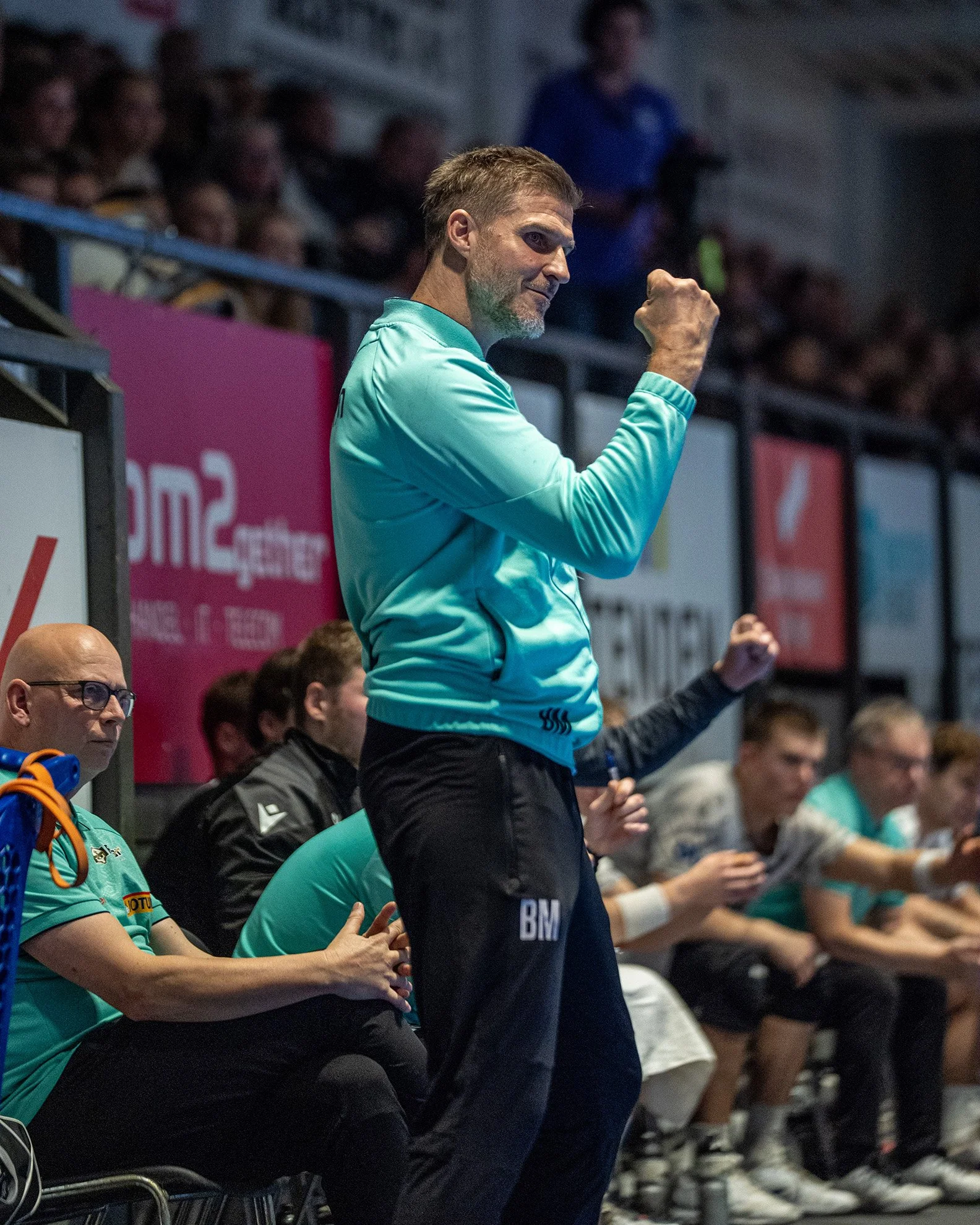 En mann iført en lys blå treningsdress jubler med knytt nevrer i en hånd mens han står ved siden av andre personer som sitte på benk i en sportshall.