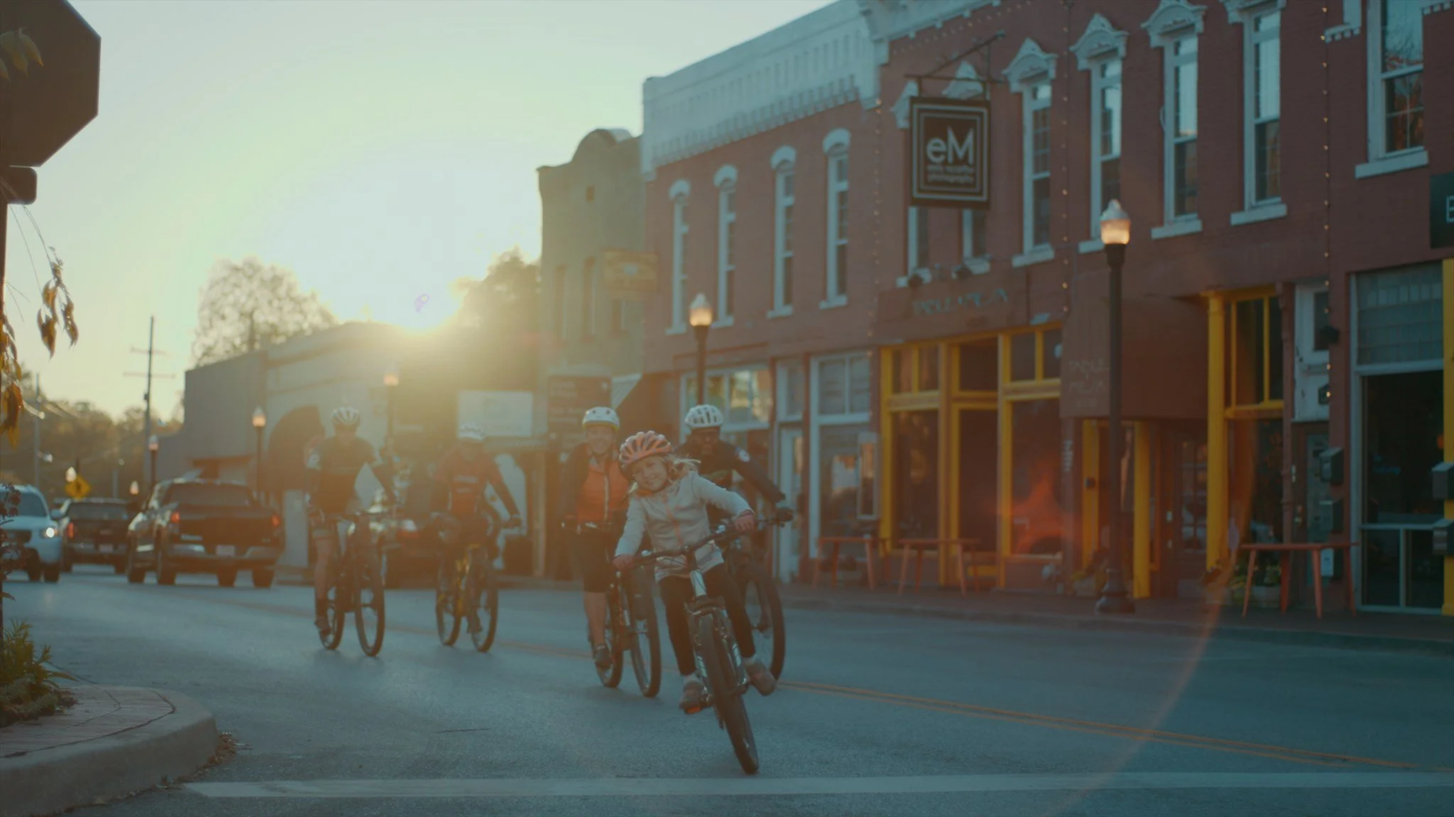 Children riding bikes on a city street at sunset, with cars parked along the road and buildings with lit windows on the sides.