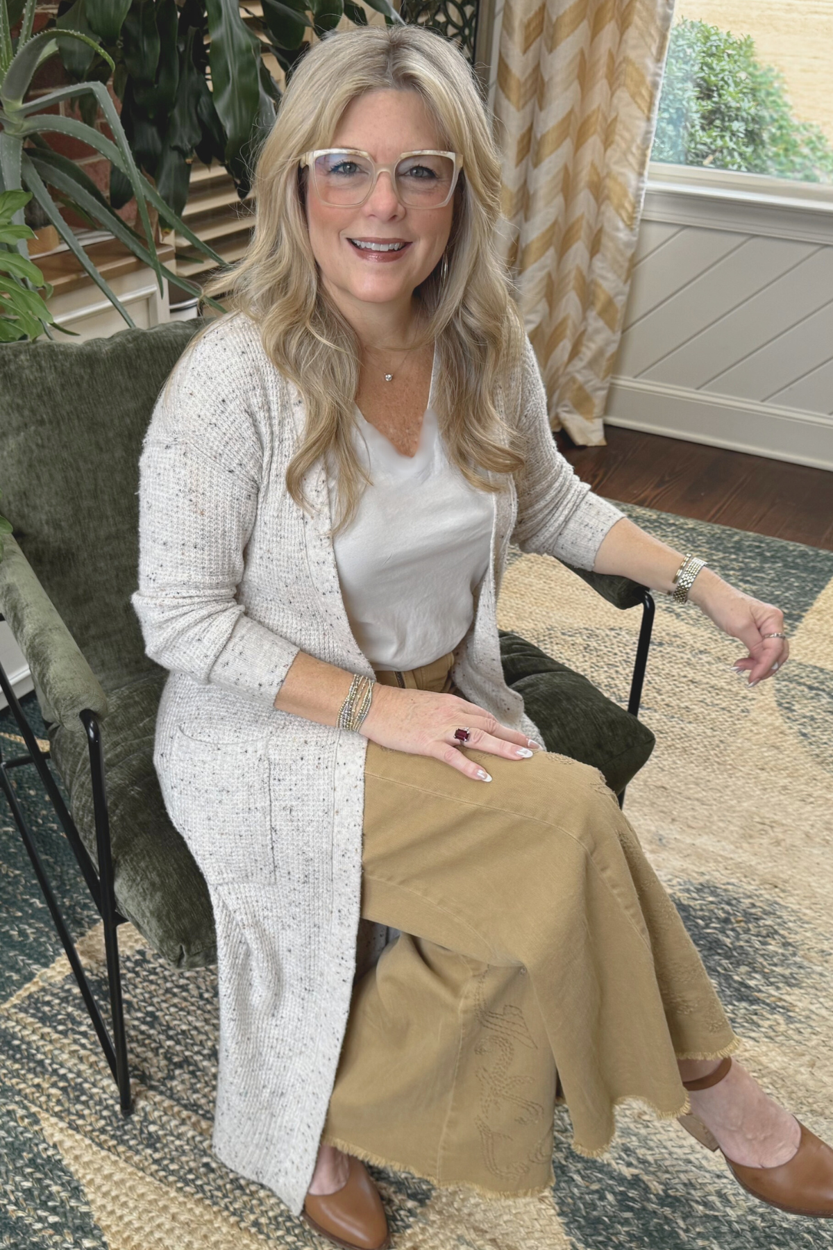 A woman with long blonde hair and glasses sitting on a green velvet armchair near a window with patterned curtains, smiling at the camera.