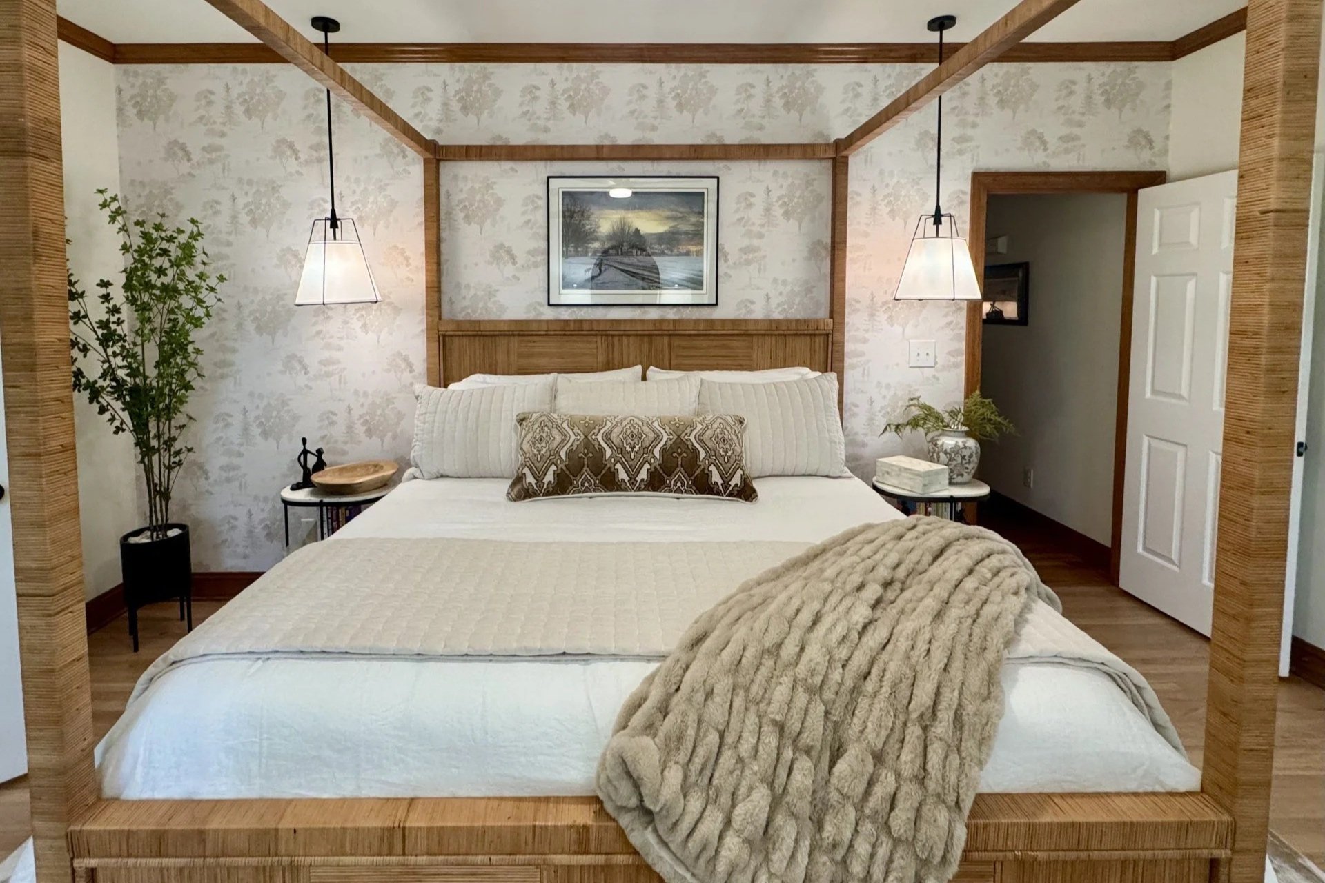 A bedroom with a wooden four-poster bed, white bedding, a textured throw pillow, a rug, a side table with books, wall art, a dresser with lamps, and a door leading outside to a porch.
