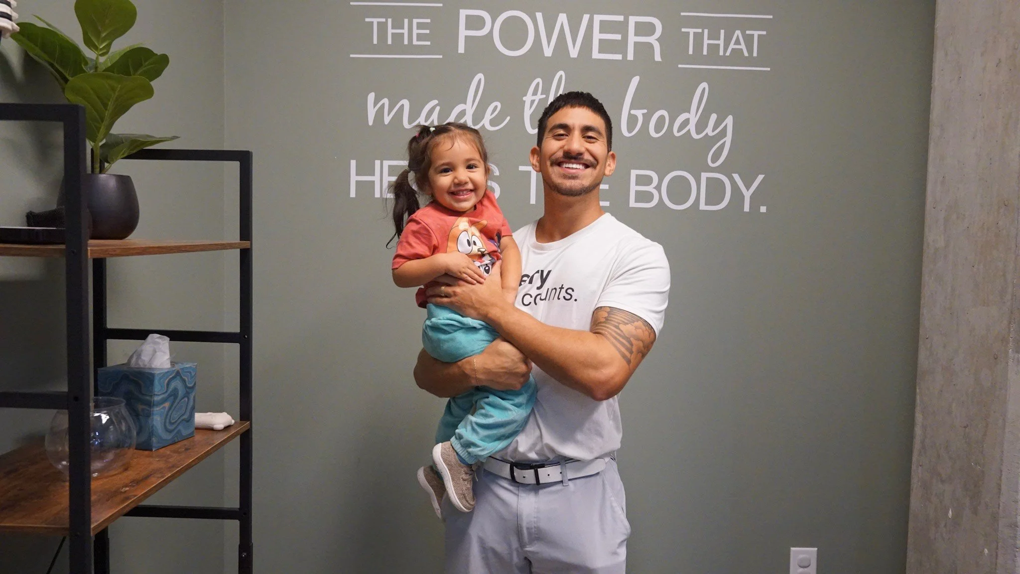 A man holding a smiling young girl indoors, with a wall behind them displaying the quote 'The power that made the body has the body.' a shelf with a plant and tissue box are also visible.