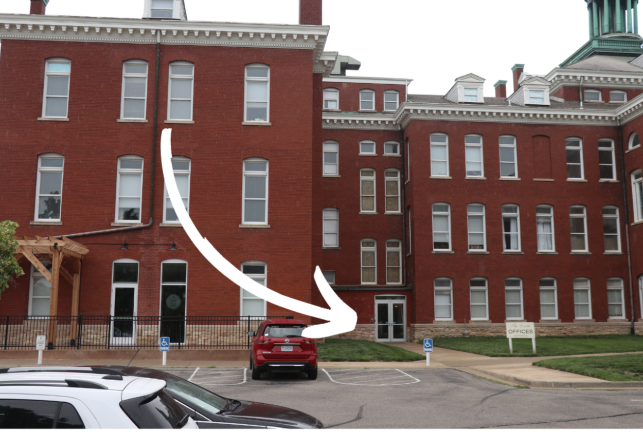 Red brick multi-story building with multiple windows, a small lawn, a parking lot with a red car, and a white sign that says 'OFFICES'.