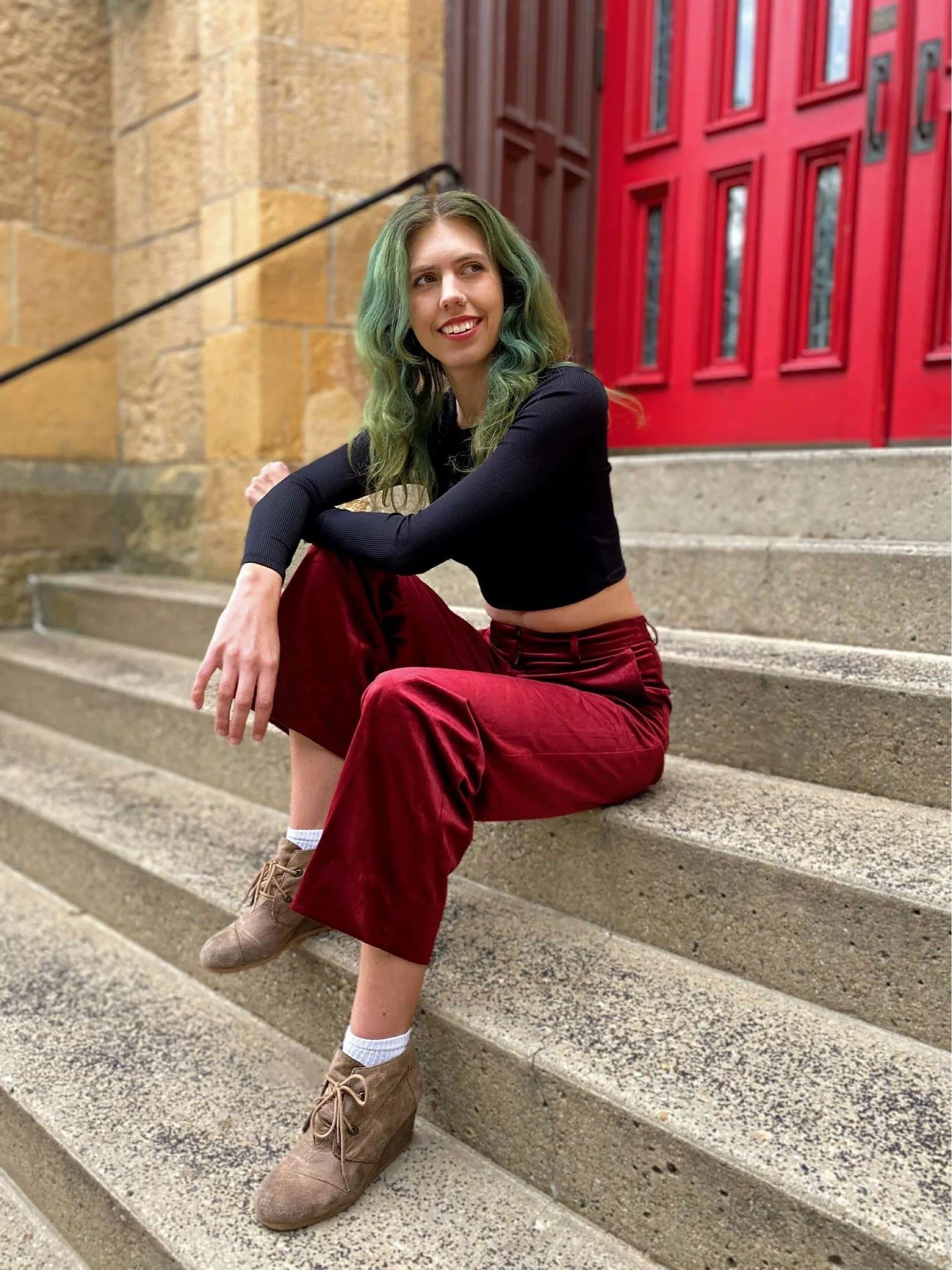 Ashley, a white non-binary person, sits on concrete stairs in front of a red door, looking off to the right and smiling. They have long green hair, a black long sleeve top, and maroon pants.