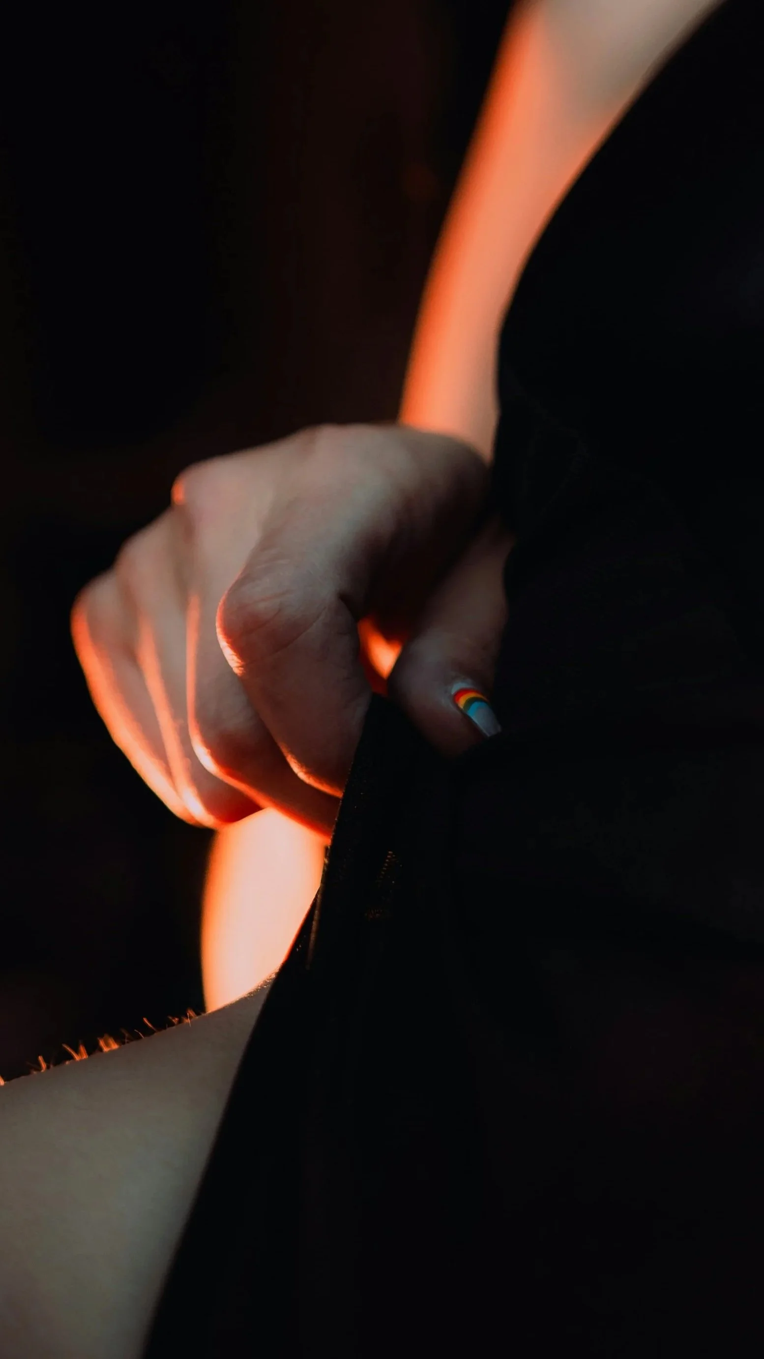 A moody close up of a hand with a rainbow painted on the thumb nail. They are lifting up their shirt, exposing the curve of their waist.