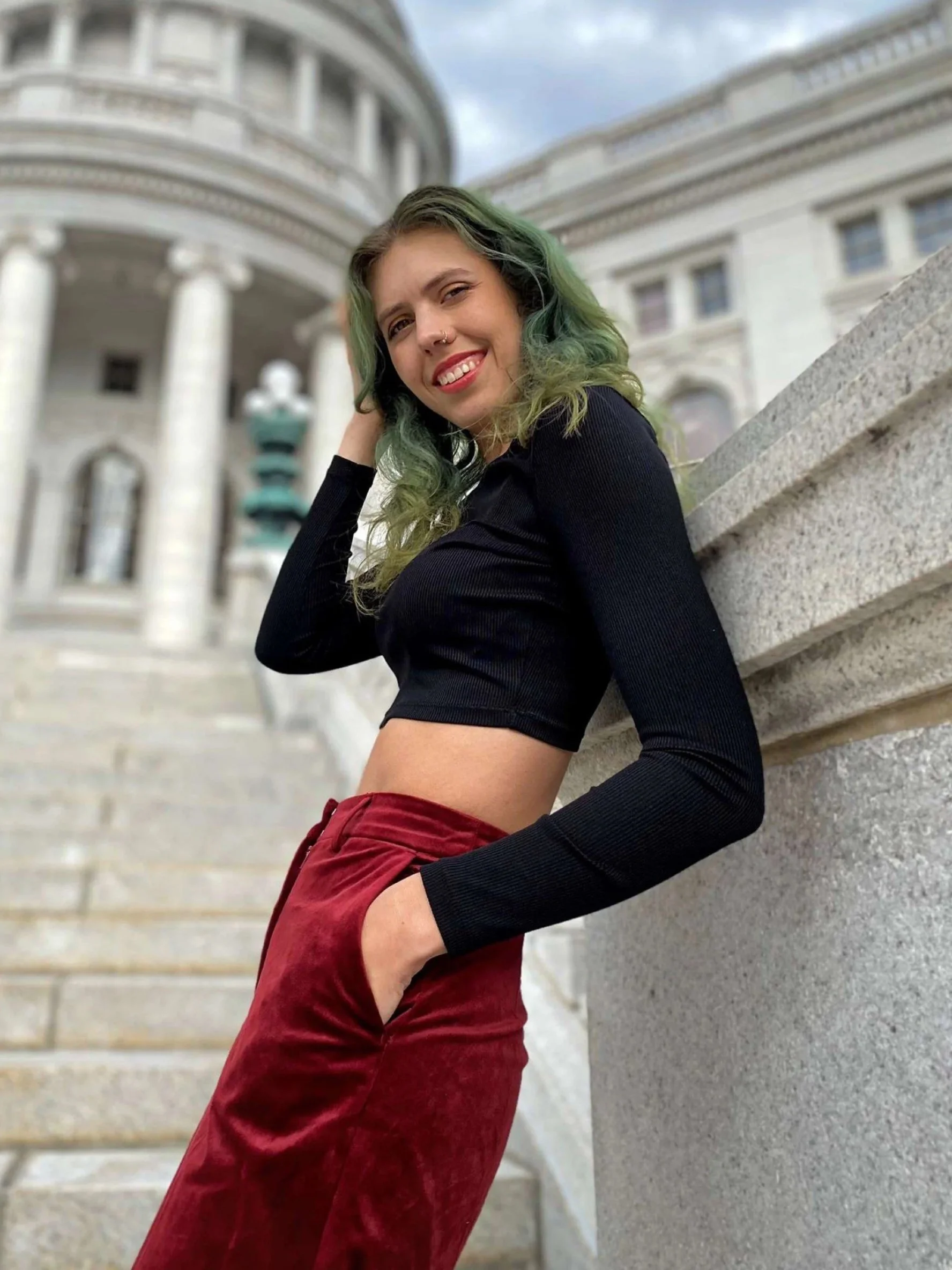 Ashley, a white non-binary person, smiles at the camera and stands on the steps of the capitol building in Madison, Wisconsin. She has long green hair, a black long sleeve top, and maroon pants.