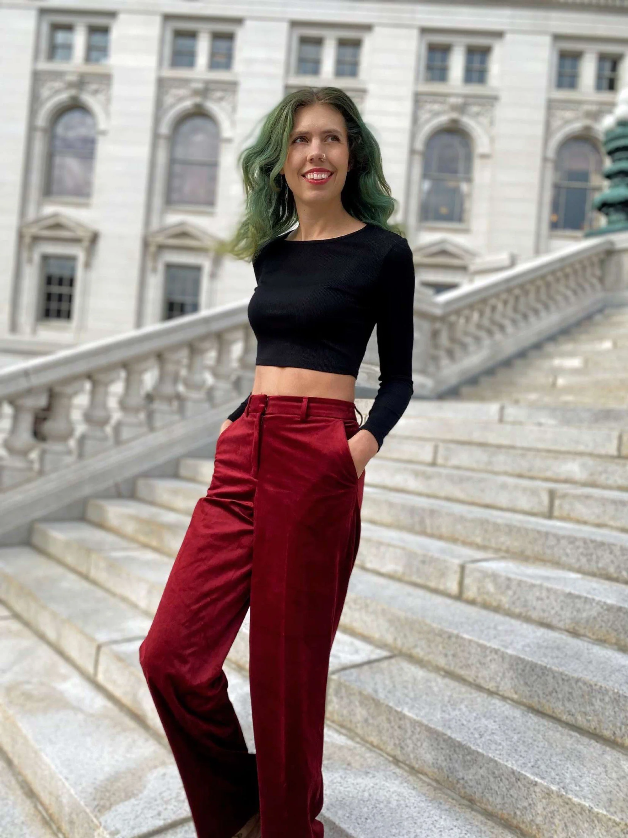 Ashley, a white nonbinary person, poses on the steps of the Madison, Wisconsin capitol building, looking to the right and smiling. They have long green hair, a black long sleeve top, and maroon pants.