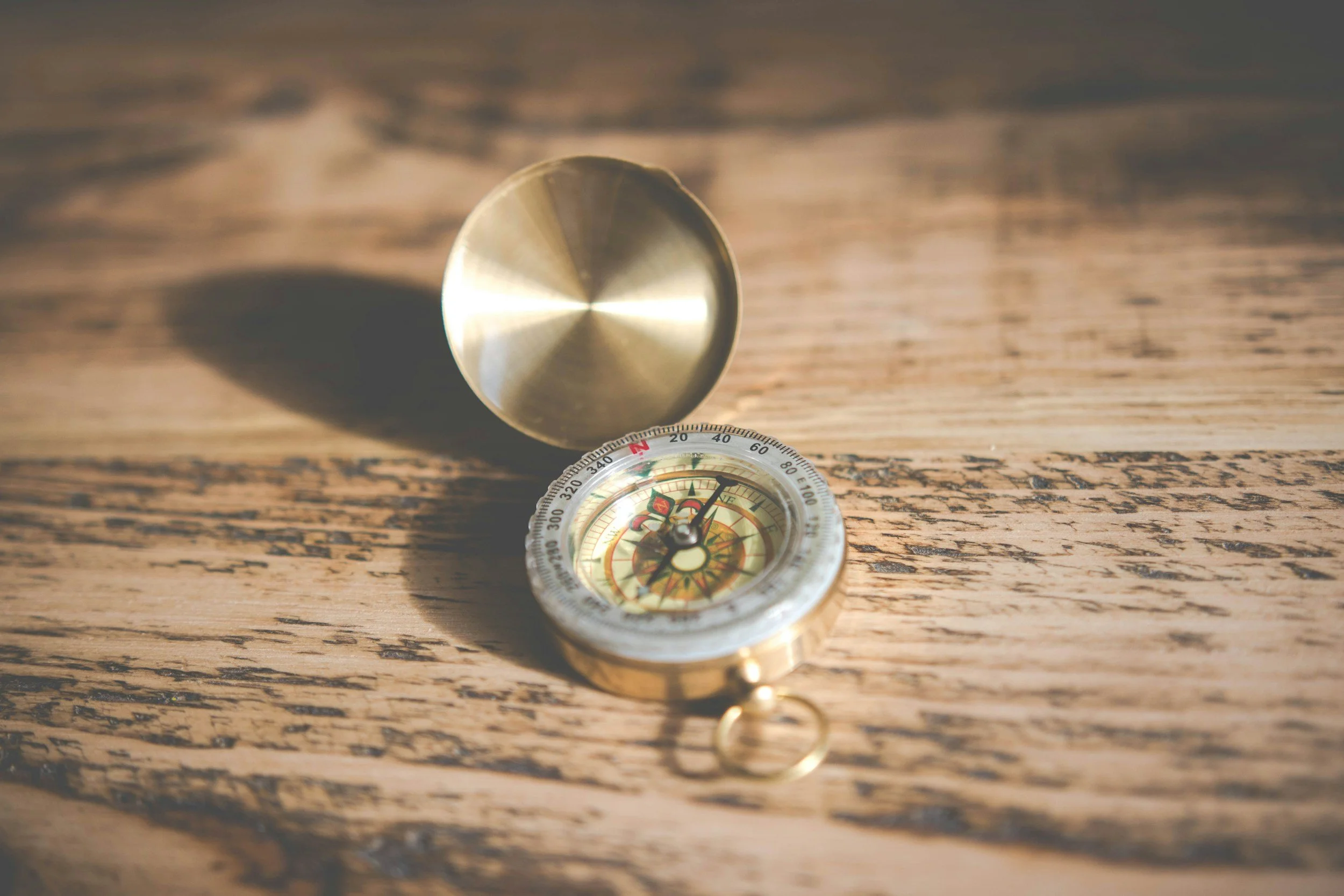 A compass with a shiny metallic cover, lying open on a wooden surface. The compass face shows cardinal directions and needle pointing north, casting a shadow on the surface.