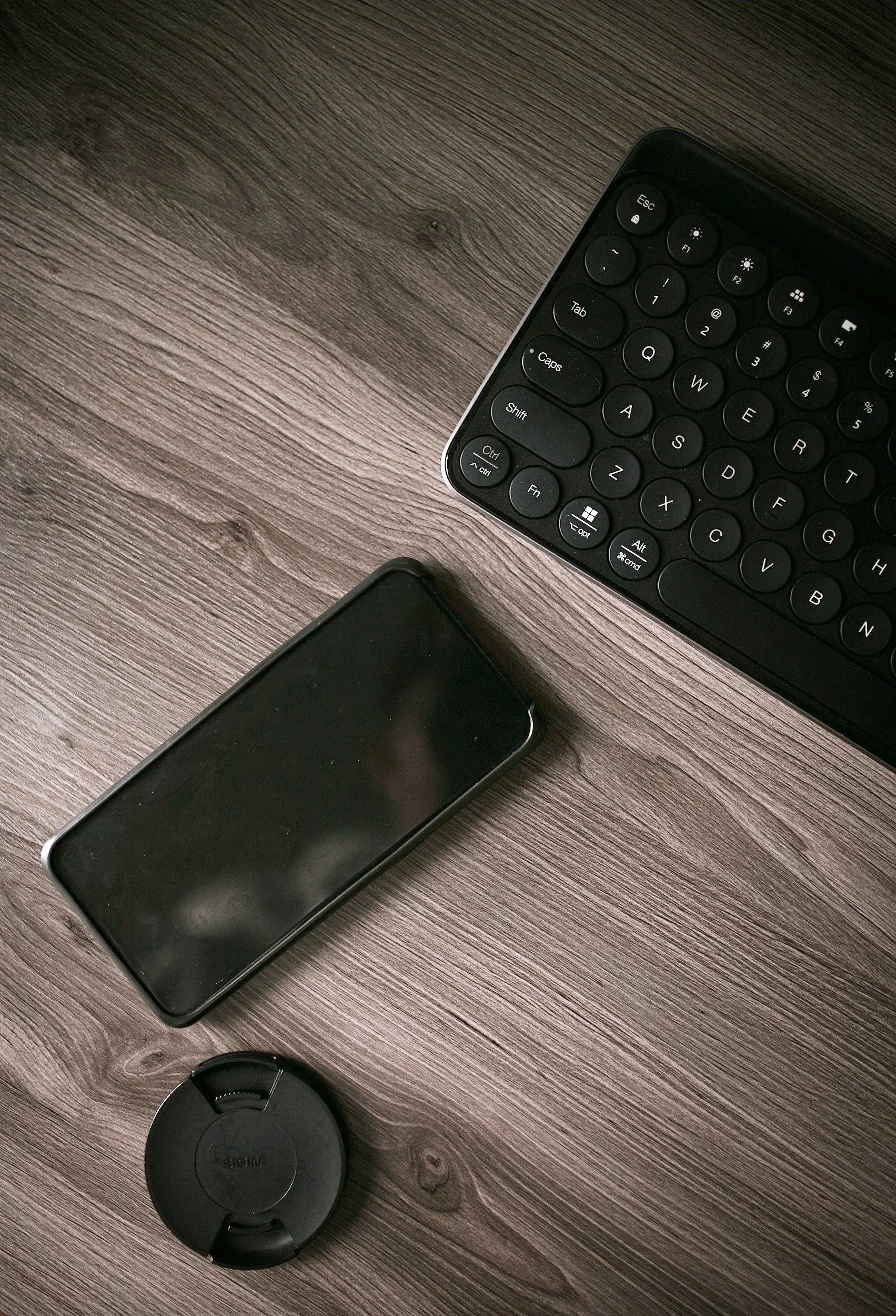 Close-up of a wireless keyboard, smartphone, and camera lens on a wooden surface.