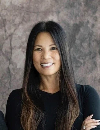 A smiling Filipina woman with long black hair wearing a black dress, standing in a studio.