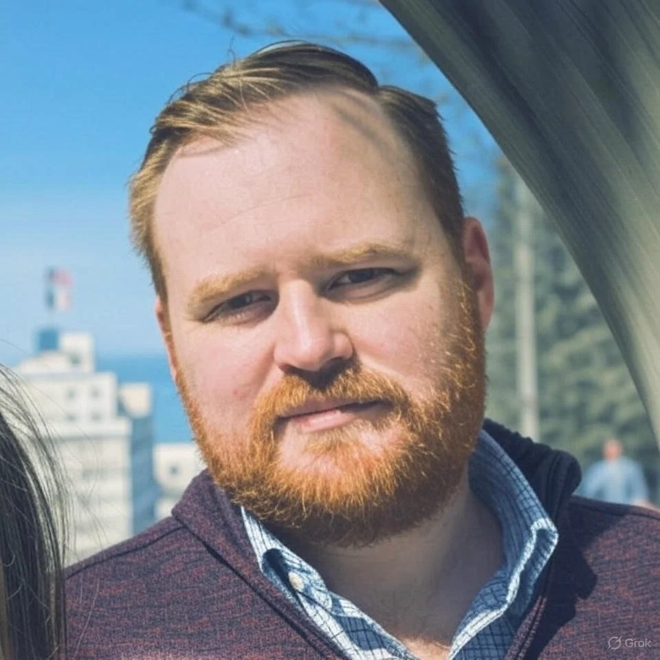 A man with light brown hair, with a beard, and wearing a collared shirt standing outdoors