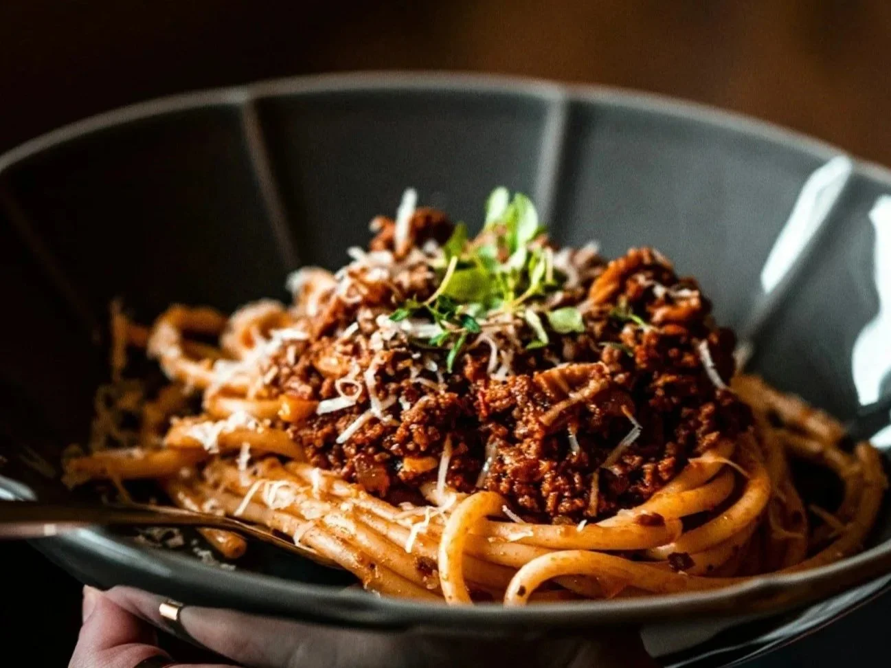A black bowl of spaghetti topped with meat sauce, shredded cheese, and fresh herbs.