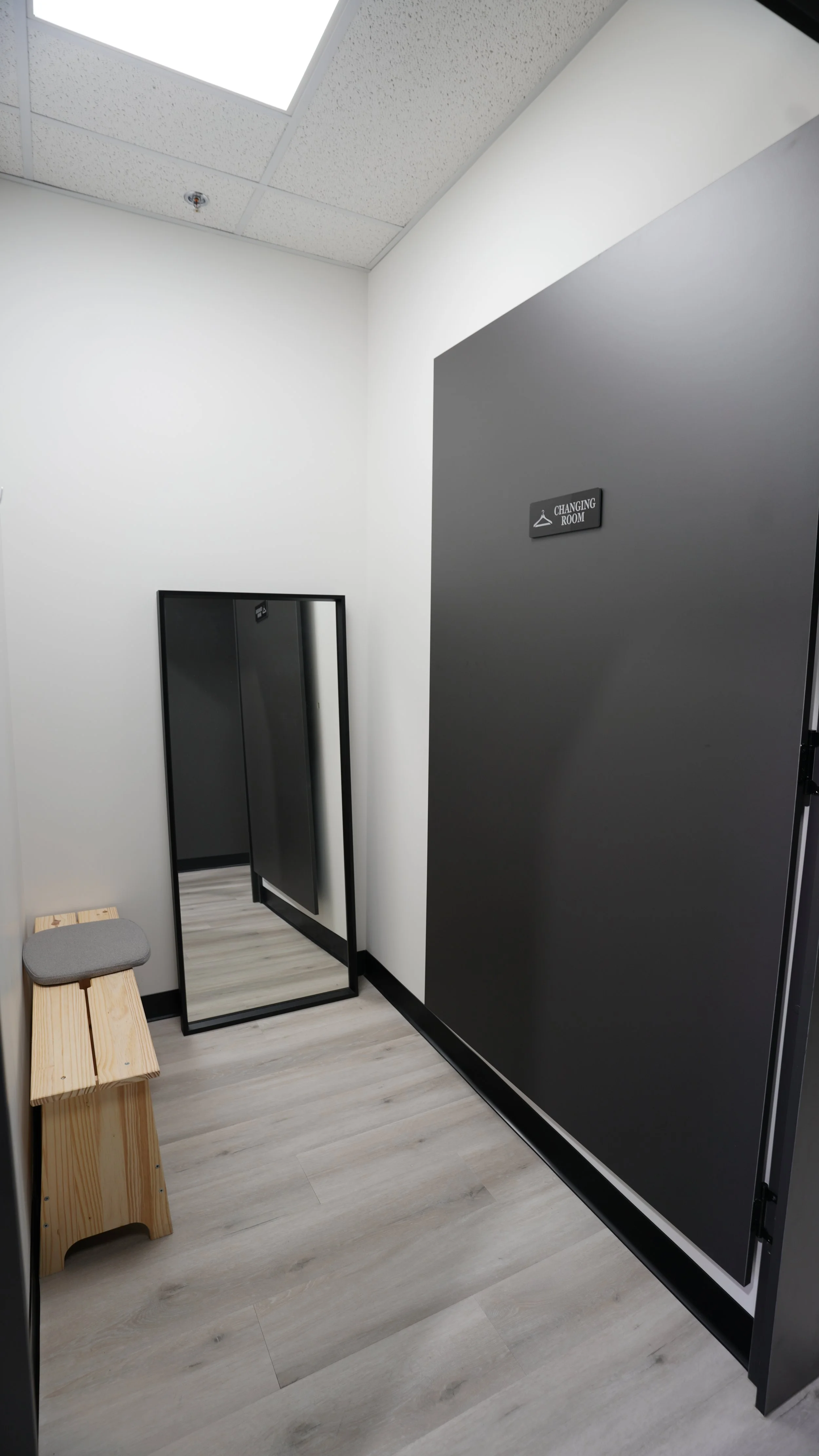 A small dressing room or changing room with a bench and a full-length mirror, a door labeled 'Changing Room,' and white walls and ceiling.