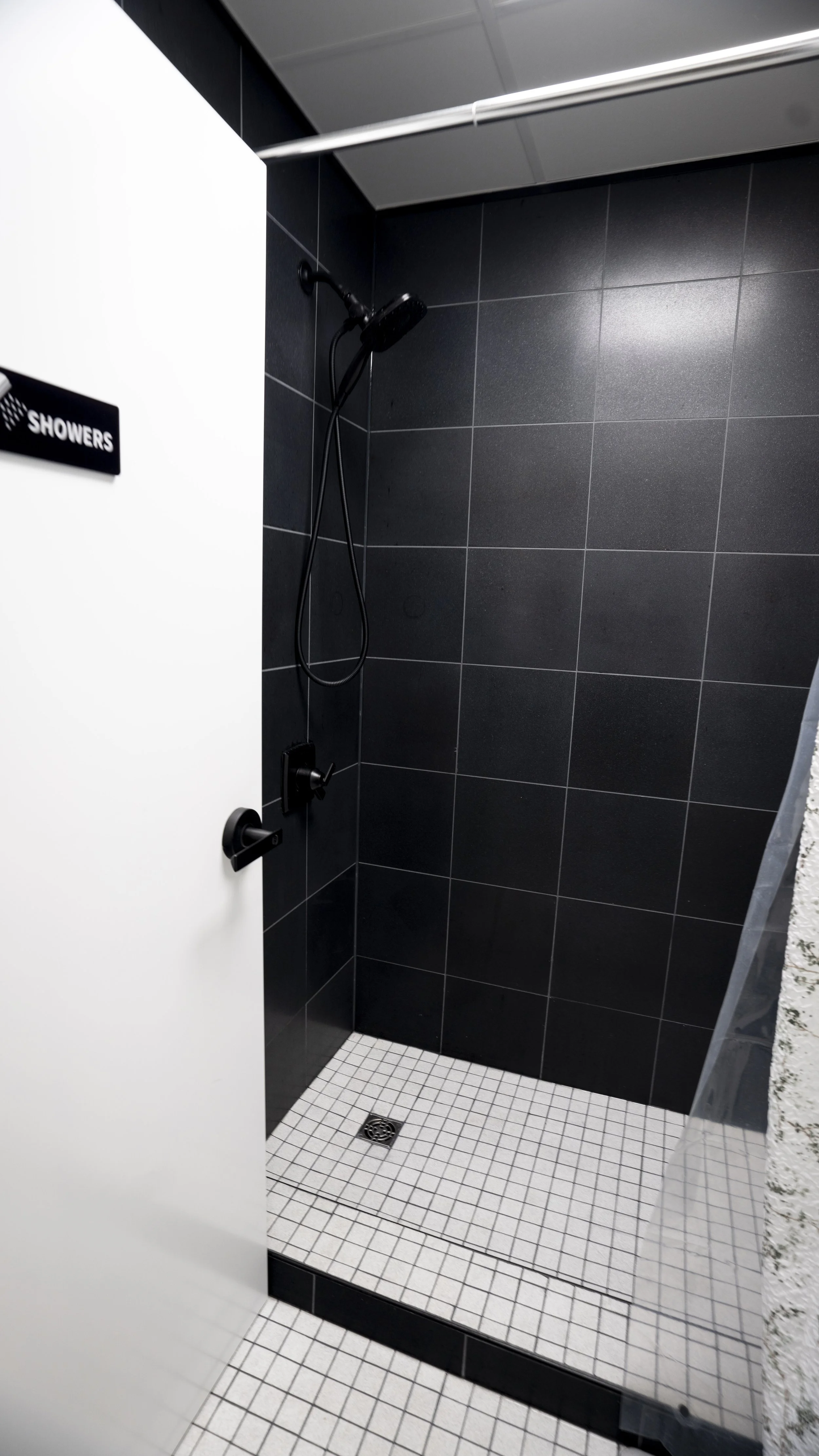 A modern shower stall with black tiled walls, a black showerhead, and a white door with a sign reading 'Showers'.