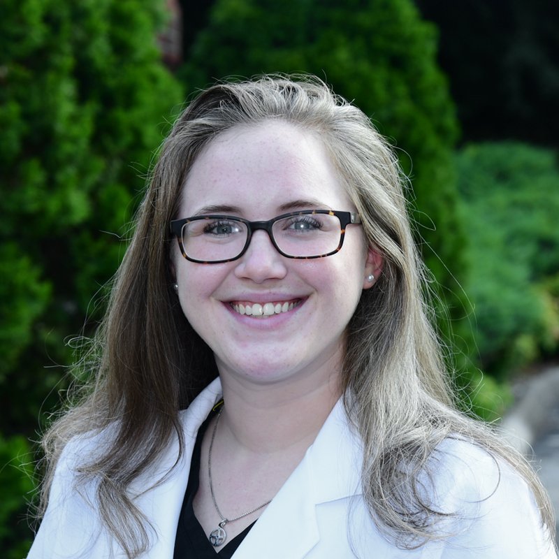 A young woman with long light brown hair, glasses, and earrings smiling outdoors with green trees in the background.