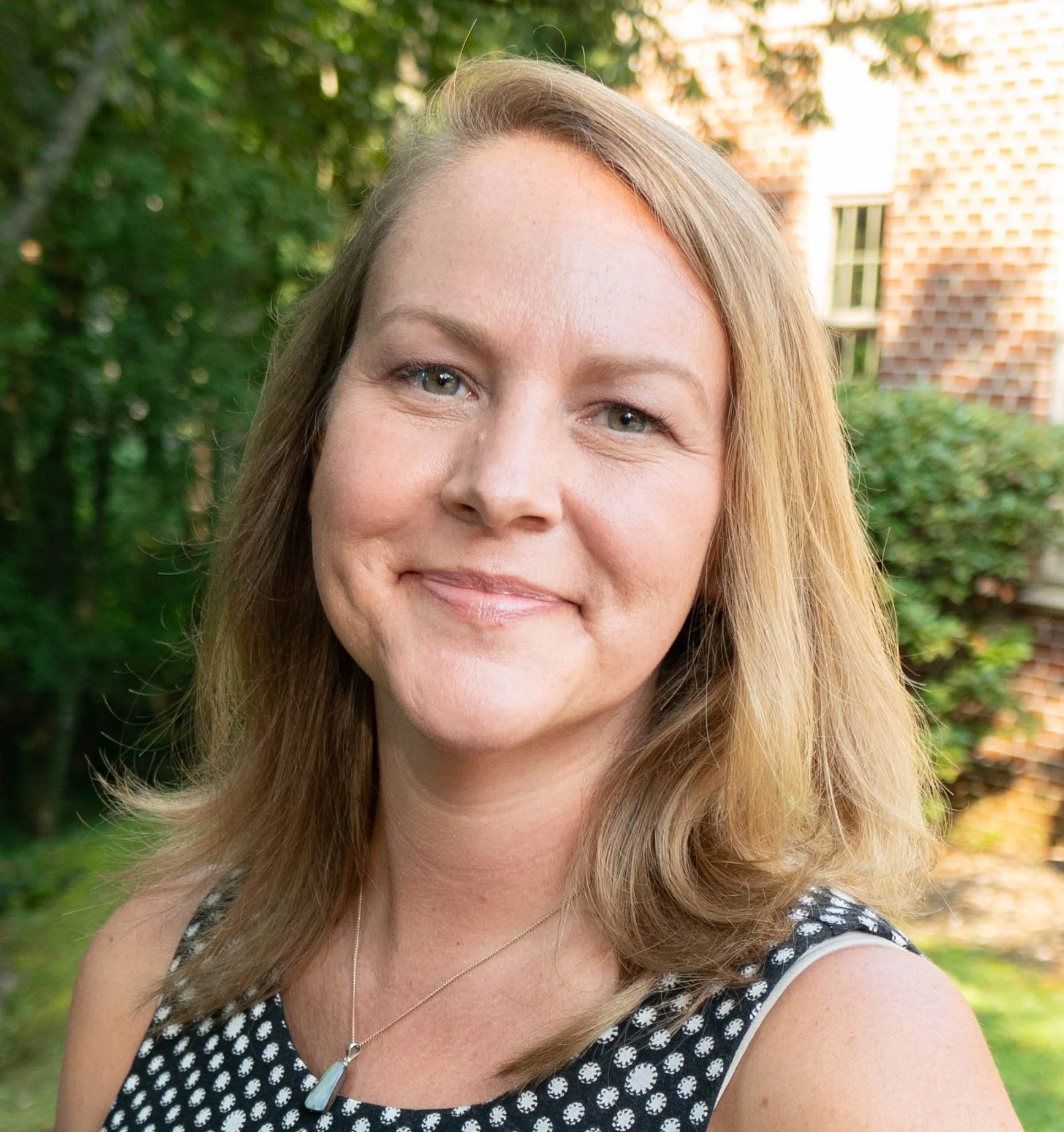 A woman with shoulder-length light brown hair, blue eyes, and fair skin smiling outdoors with green trees and a brick building in the background.