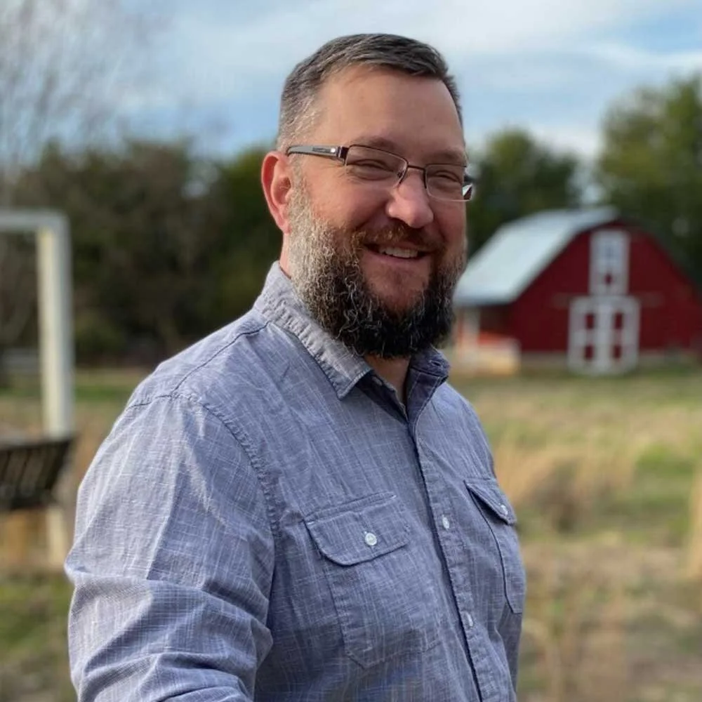 A man with glasses and a beard smiling outdoors, with a red barn in the background.
