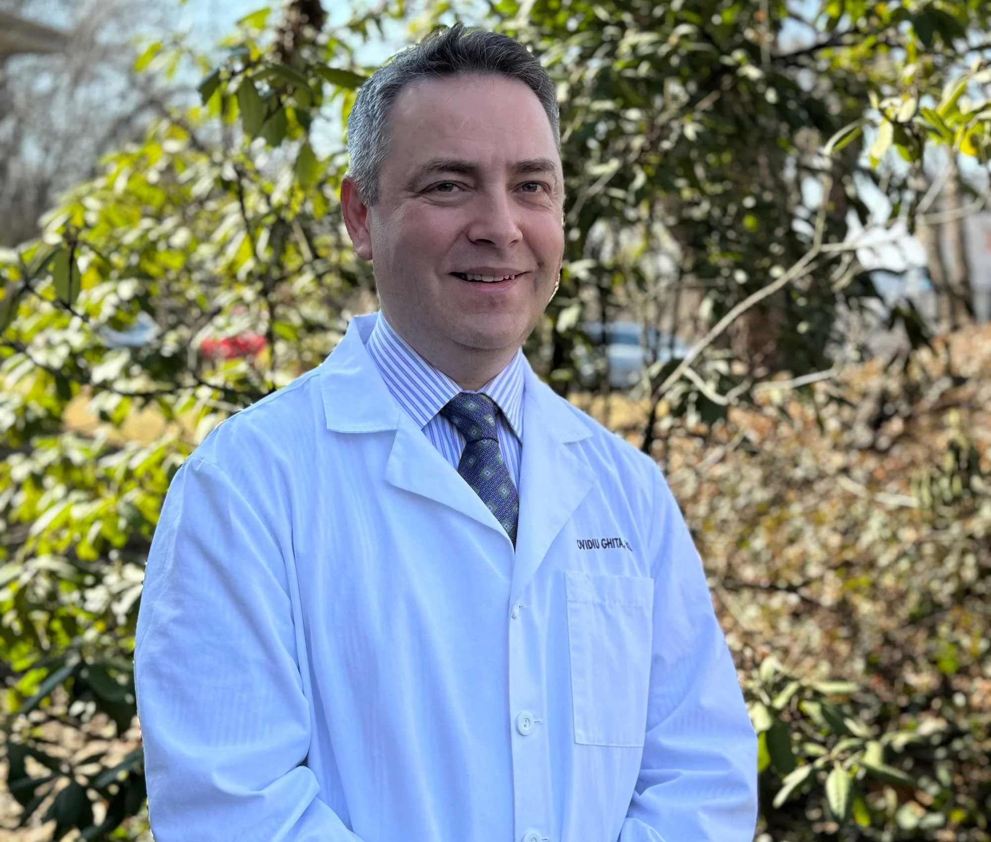 A man wearing a white coat and tie standing outdoors with greenery in the background.