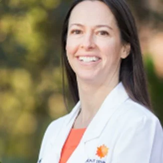 A woman smiling outdoors with trees in the background, wearing a white jacket with a logo.