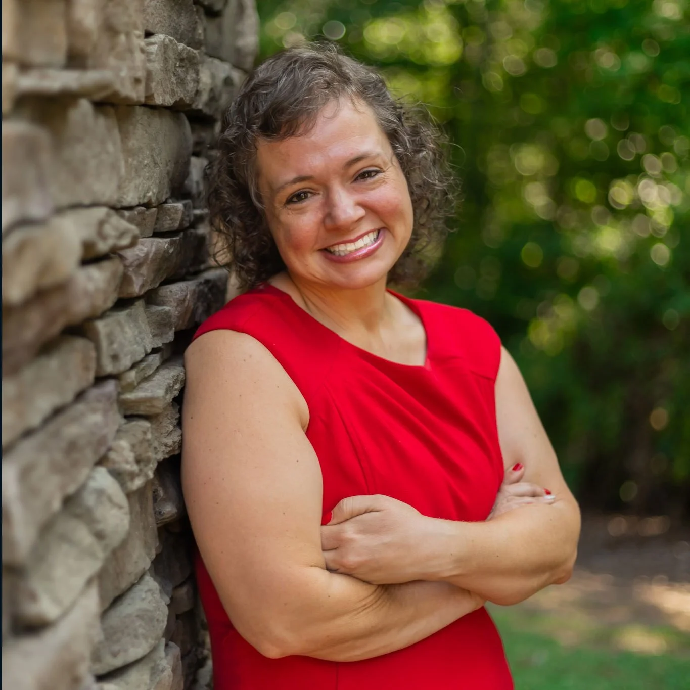 A woman with curly brown hair wearing a red sleeveless dress, smiling and standing with arms crossed next to a stone wall outdoors with green blurred foliage in the background.