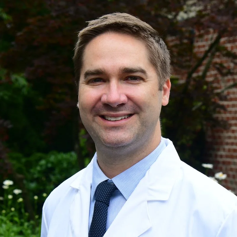 A smiling male doctor with short brown hair, dressed in a white lab coat, blue shirt, and dark blue tie, standing outdoors with green foliage and a brick building in the background.