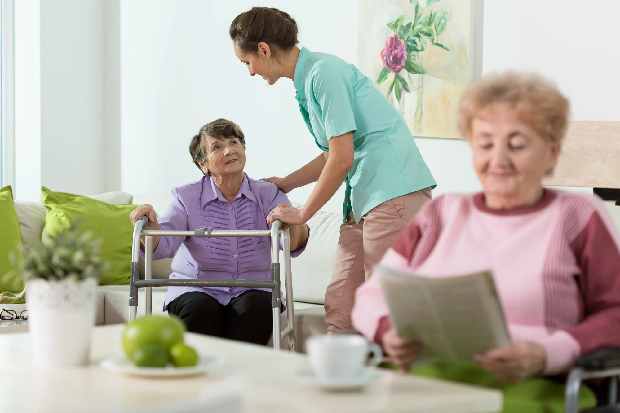 A caregiver helping an elderly woman with a walker during a visit in a living room, with another elderly woman reading a book at the table.
