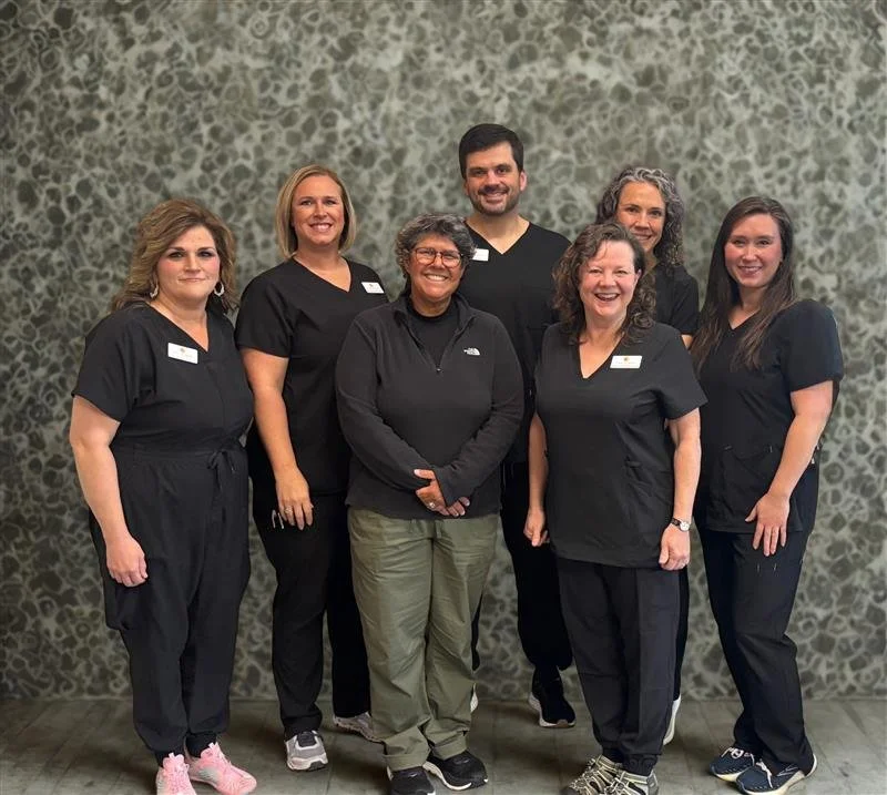 Group of seven women and one man standing together indoors in front of a patterned wall, dressed in black scrubs, smiling at the camera.