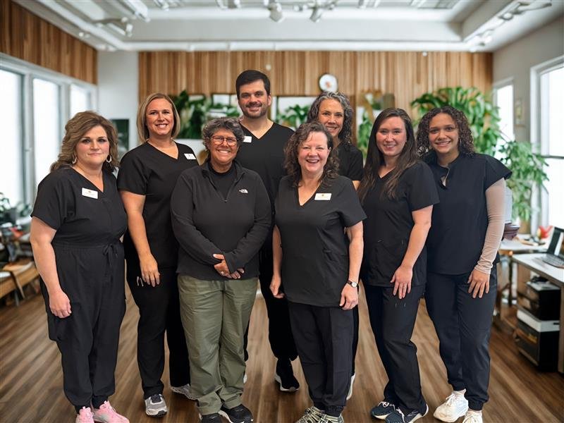 Group of eight healthcare or dental professionals standing in a well-lit room with windows and plants, smiling at the camera.