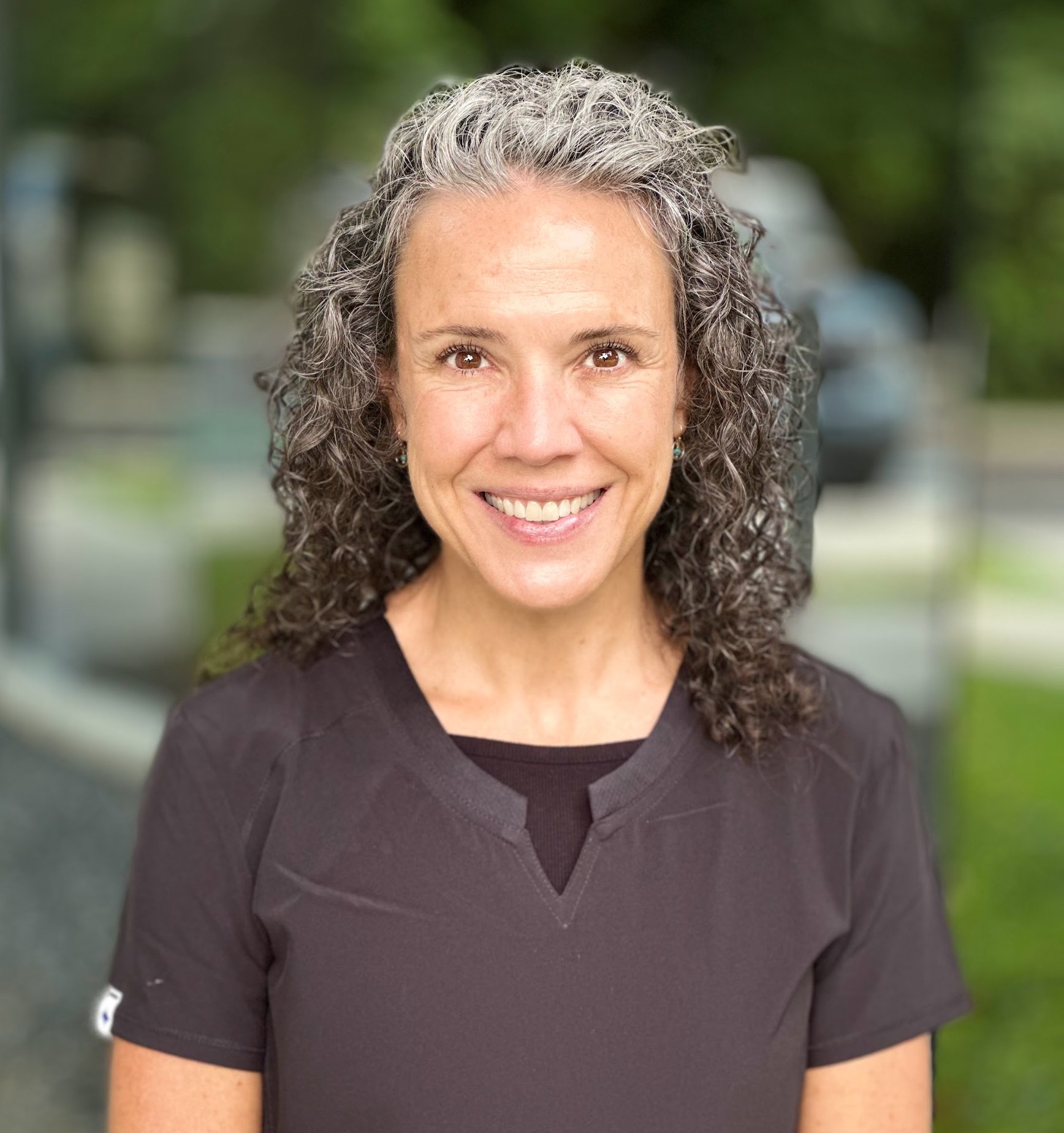 A smiling woman with curly gray hair outdoors in a blurred background.