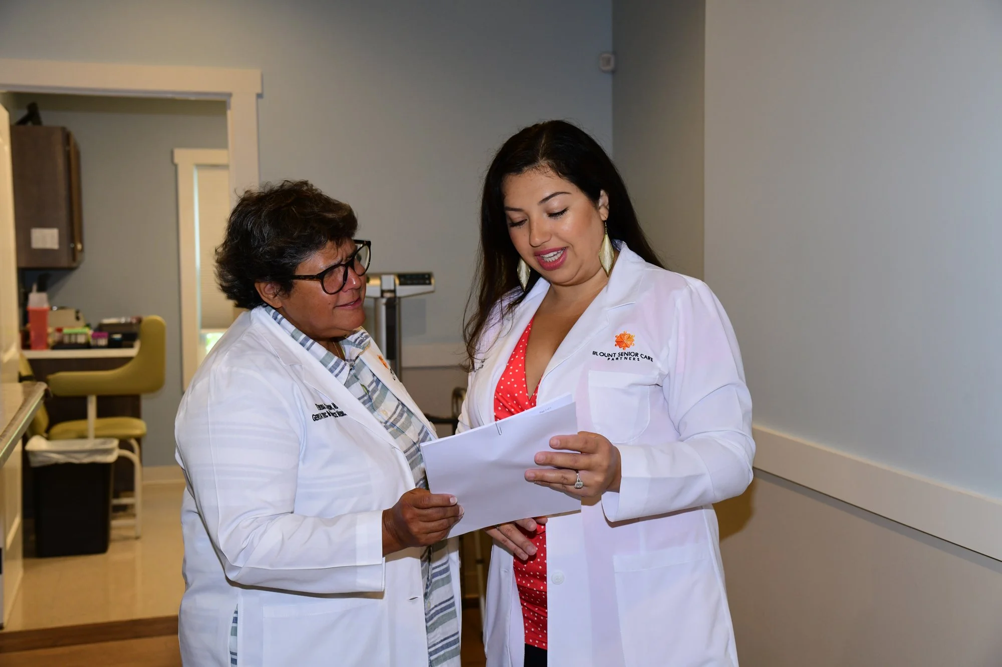 Two women in white lab coats standing in a medical facility, discussing a document.