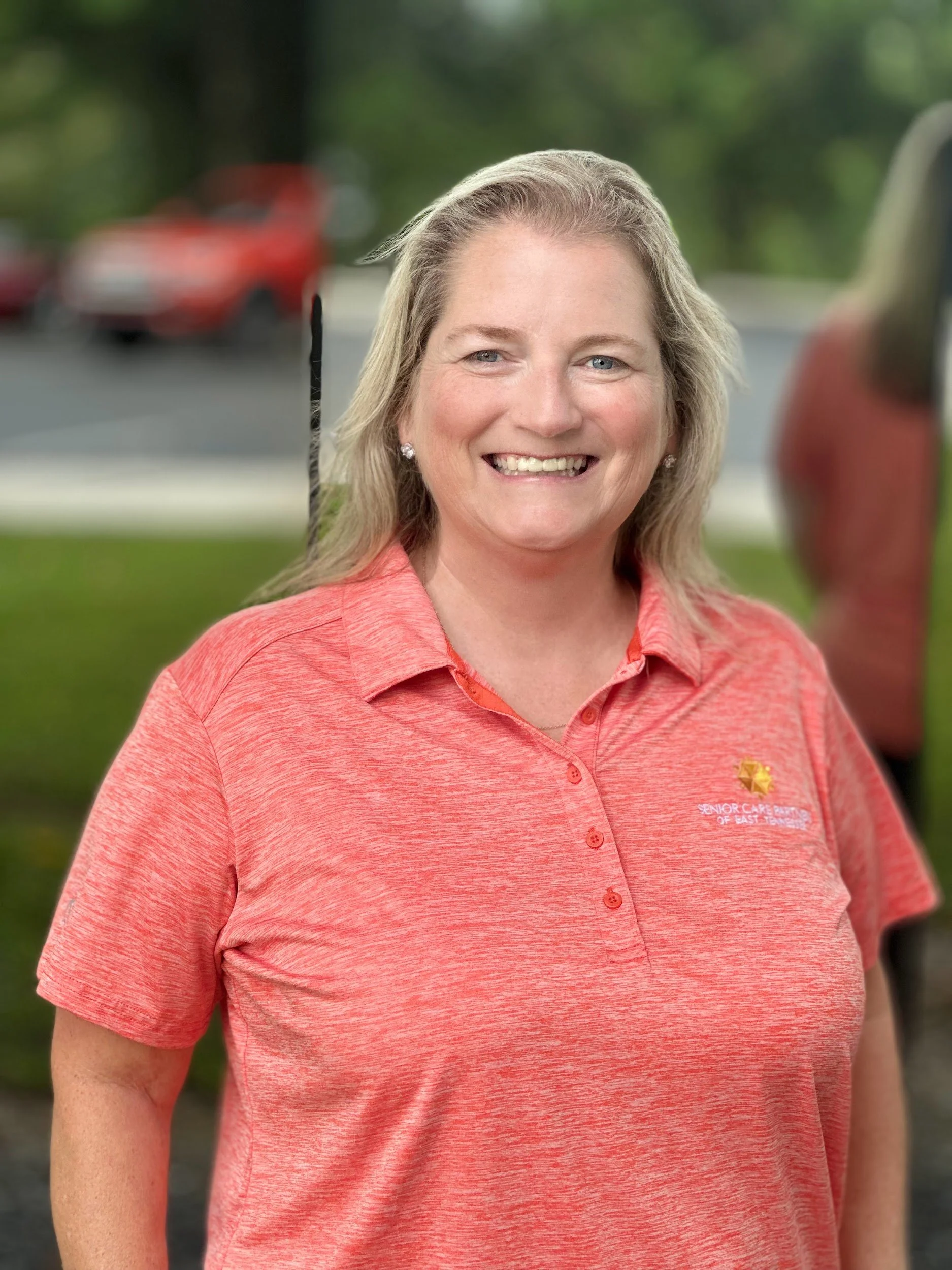 A woman with shoulder-length blonde hair smiling outdoors, wearing a pink polo shirt with a logo on the left chest that reads 'Senior Care'.