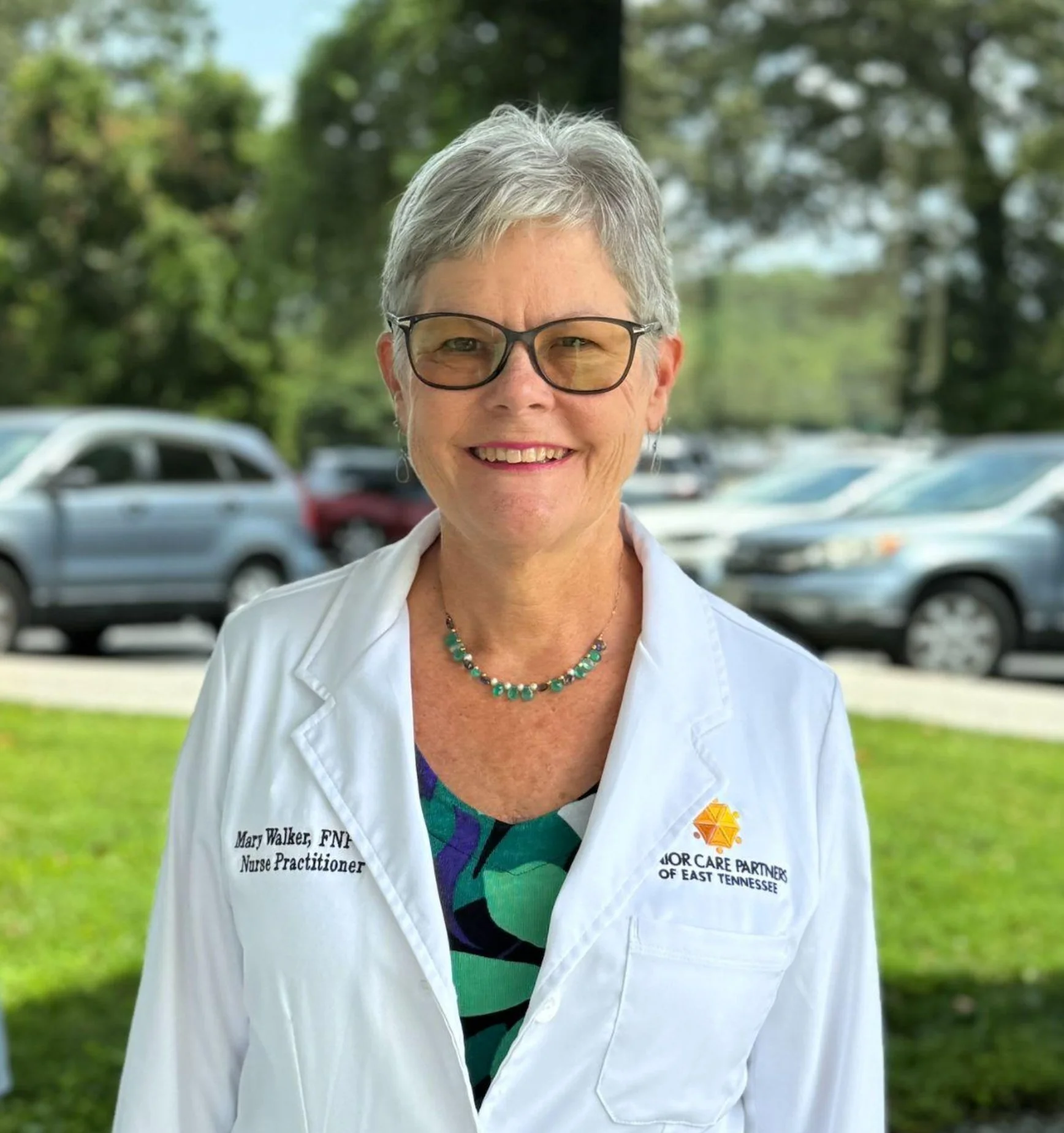 A smiling woman with short gray hair wearing glasses, a white coat with 'Mary Walker, FNP Nurse Practitioner' embroidered on it, standing outdoors in front of parked cars and green trees.