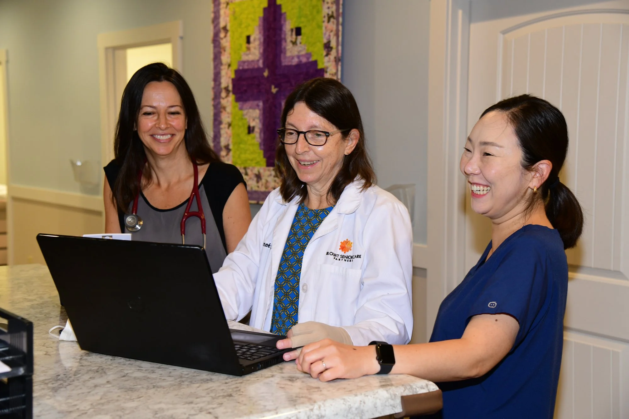 Three women, including a healthcare professional in a white coat, are gathered around a laptop in a well-lit room, smiling and looking at the screen.