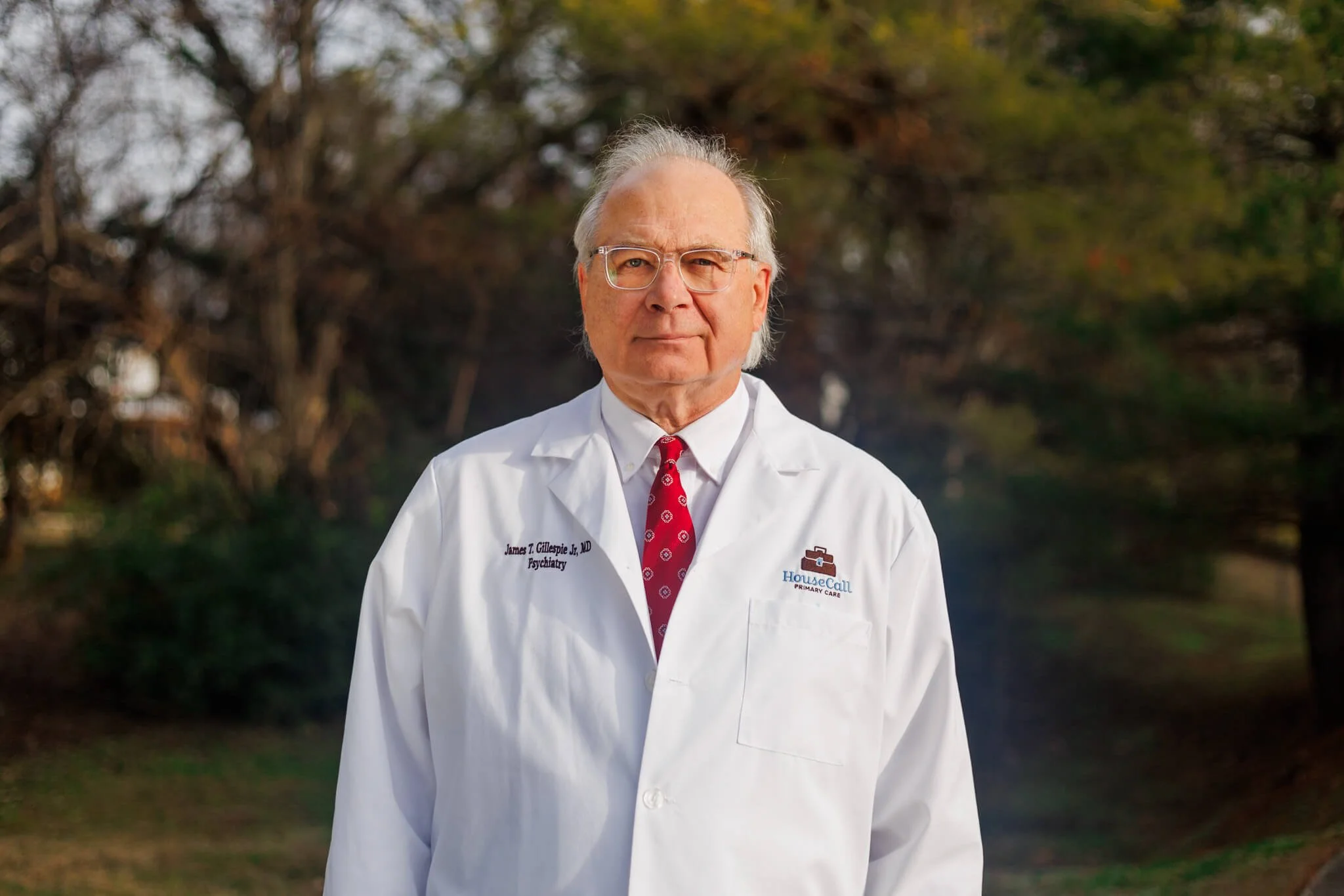 An elderly man wearing glasses, a white medical coat, and a red tie stands outdoors with trees in the background.