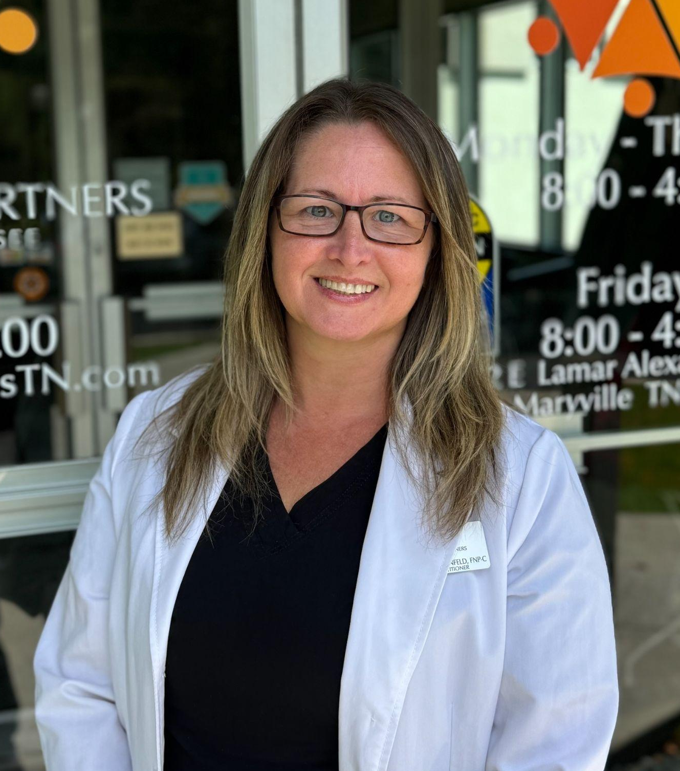 A woman with glasses, smiling, wearing a white coat, standing outside a building with a glass door.