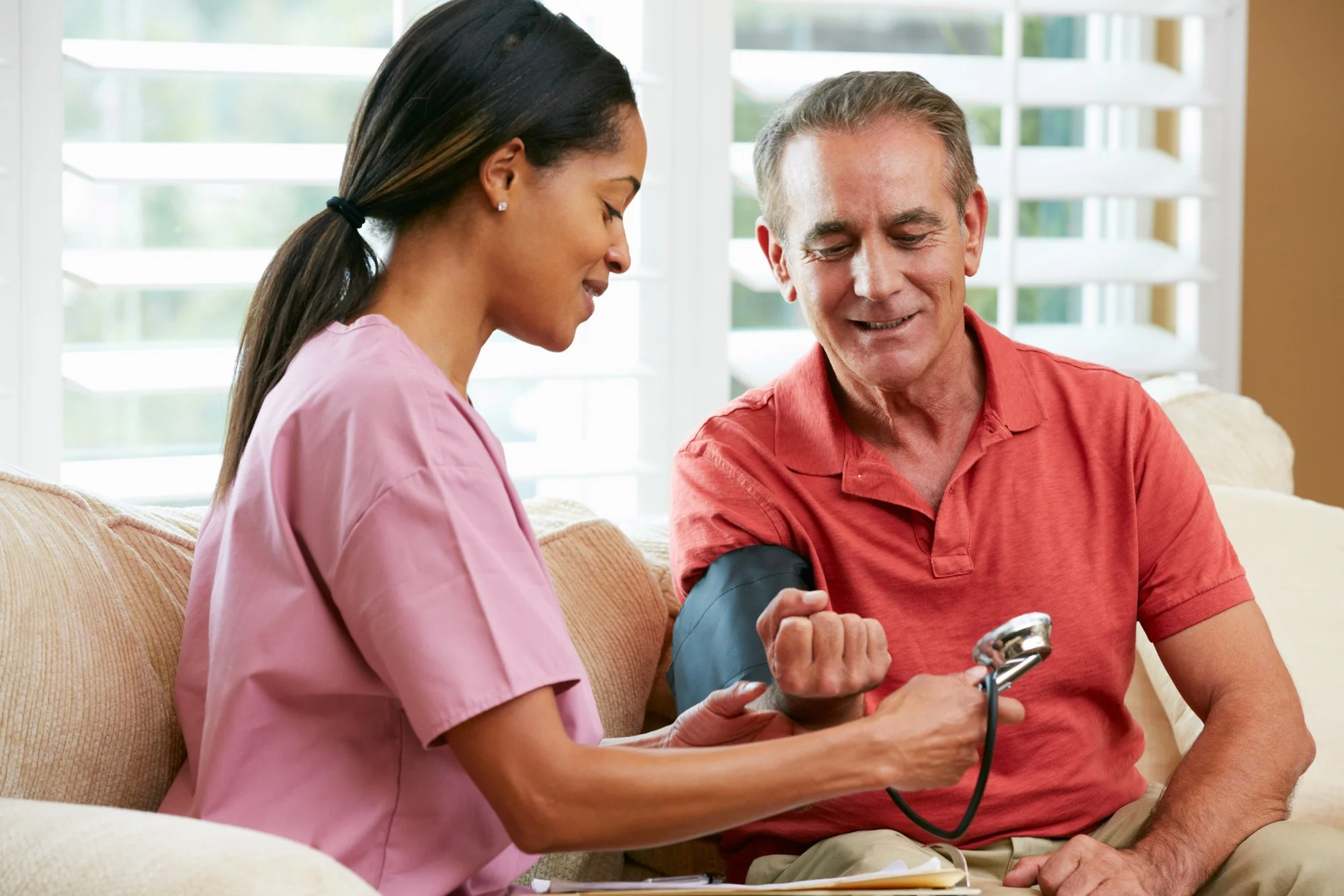 A healthcare worker in pink scrubs measuring an elderly man's blood pressure using a cuff and sphygmomanometer in a bright room with large windows.