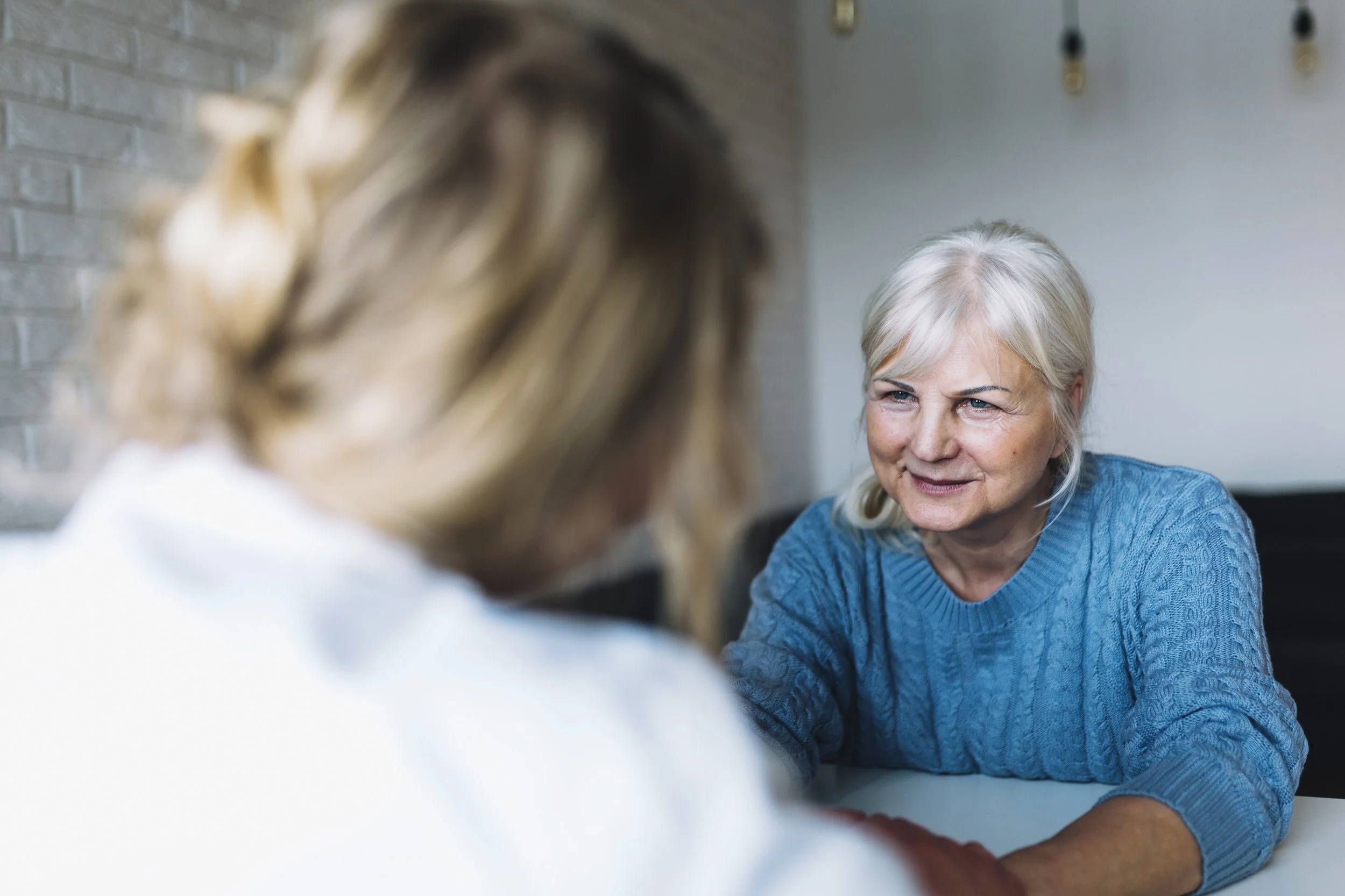An elderly woman with white hair wearing a blue sweater, smiling and talking with another person with blond hair, blurred in the foreground, indoor setting with brick wall and light fixtures in the background.