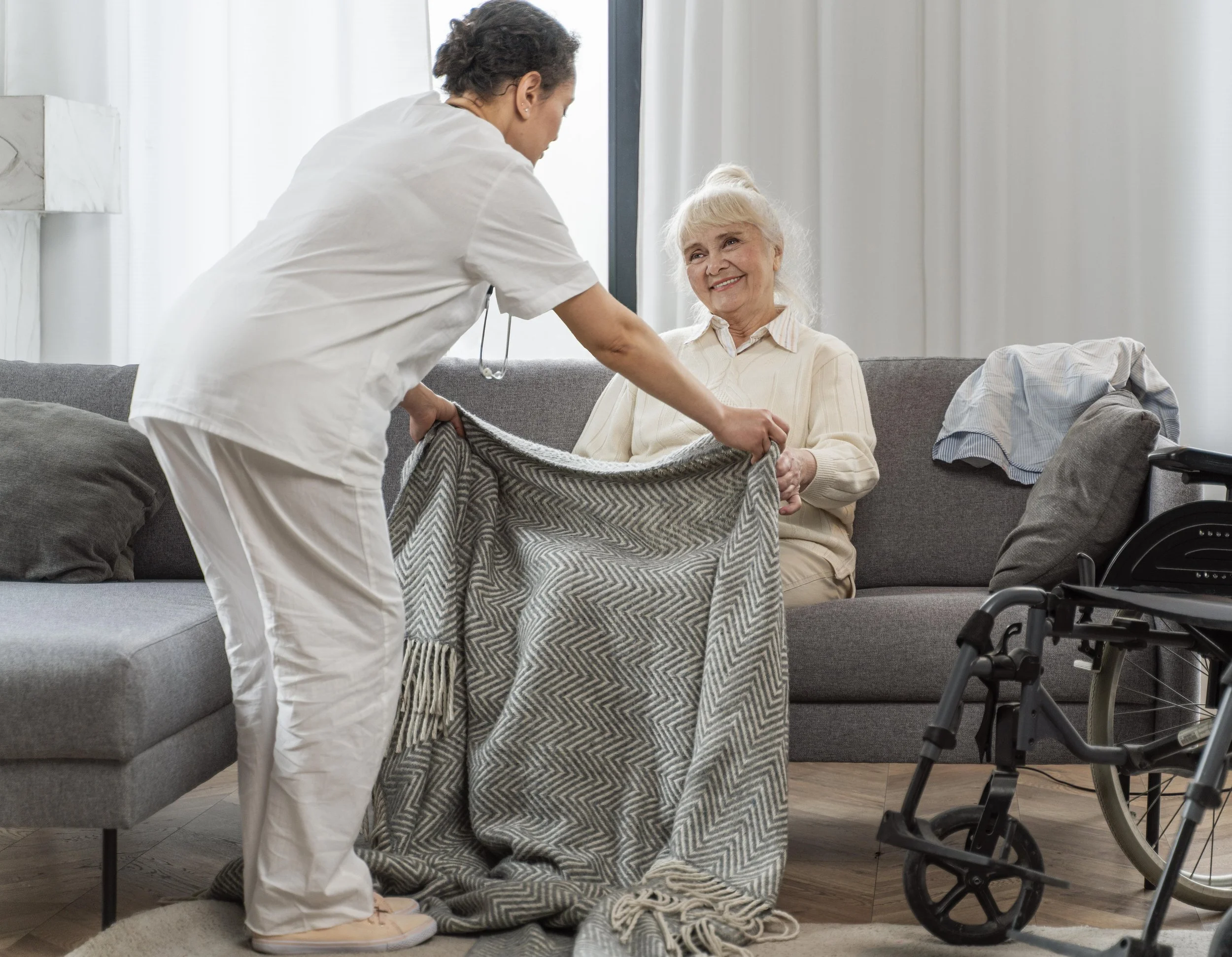 A caregiver helping an elderly woman sitting on a couch with a blanket, with a wheelchair nearby.