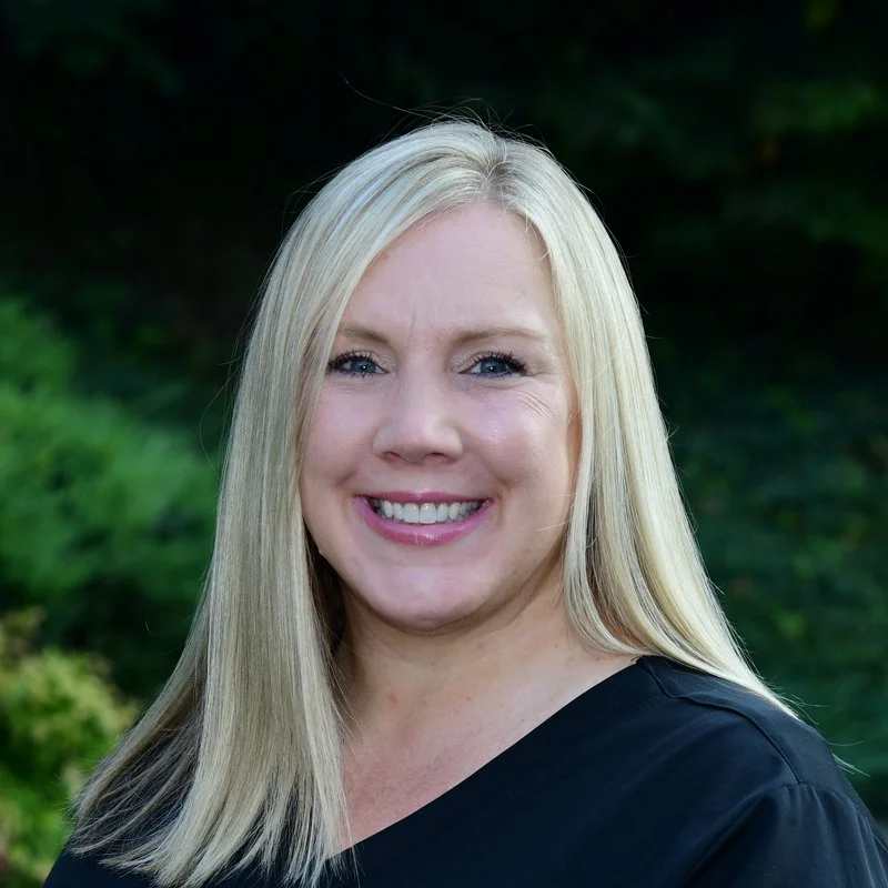 A woman with long blonde hair smiling outdoors with greenery in the background.