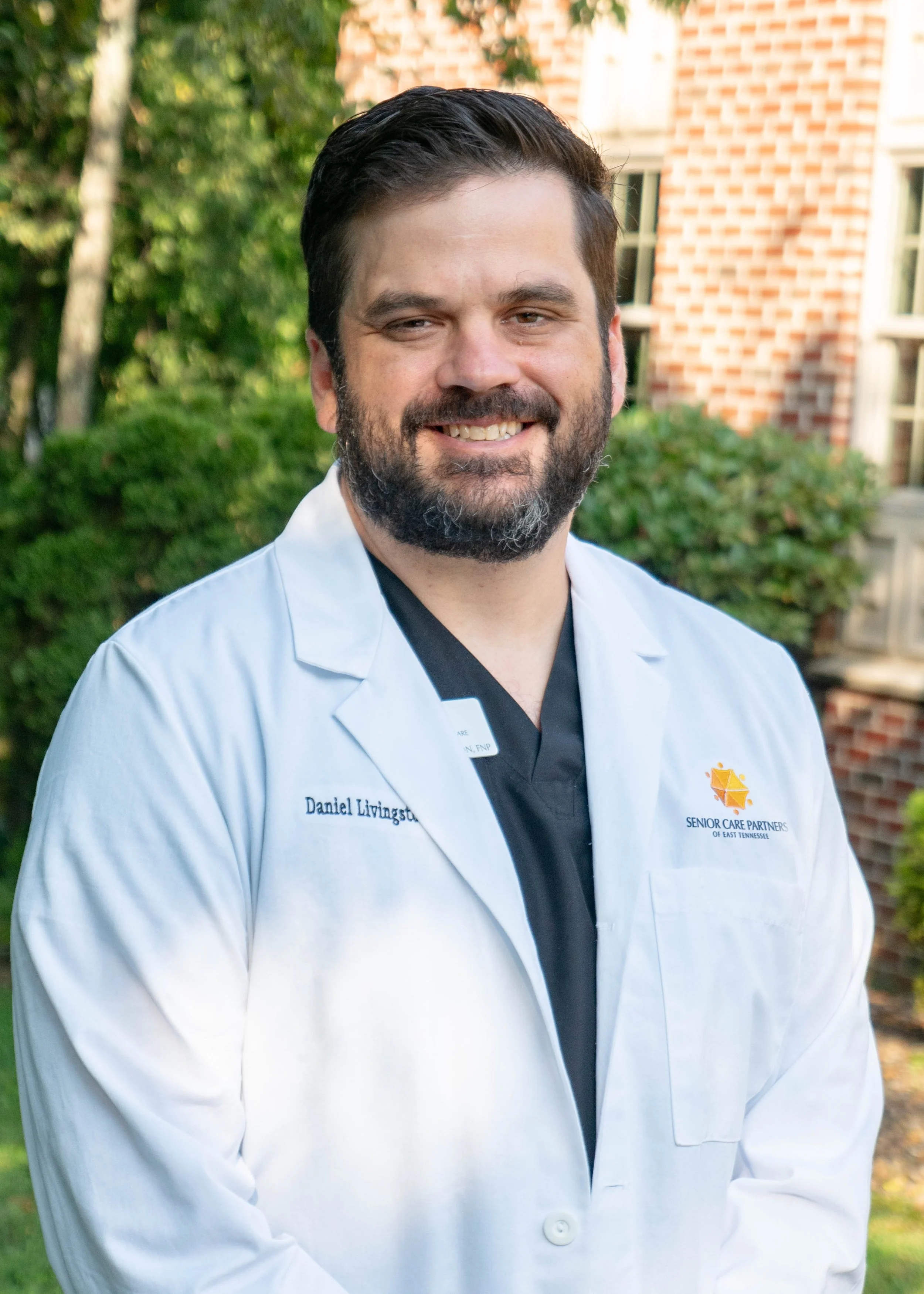 A man with dark hair and beard smiling outdoors, wearing a white medical coat with the name 'Daniel Livingstone' and a patch that says 'Senior Care Partners of East Tennessee'.