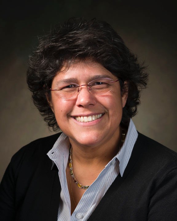 Close-up of a smiling woman with dark curly hair, wearing a light blue collared shirt and white coat, outdoors with green foliage in the background.