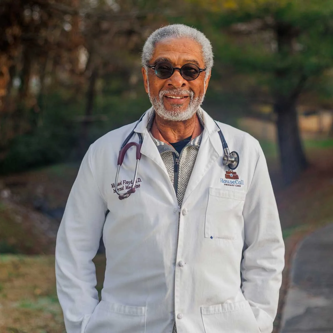 An older man with gray hair and beard, wearing sunglasses, a white medical coat with a stethoscope around his neck, smiling outdoors in a park-like setting.