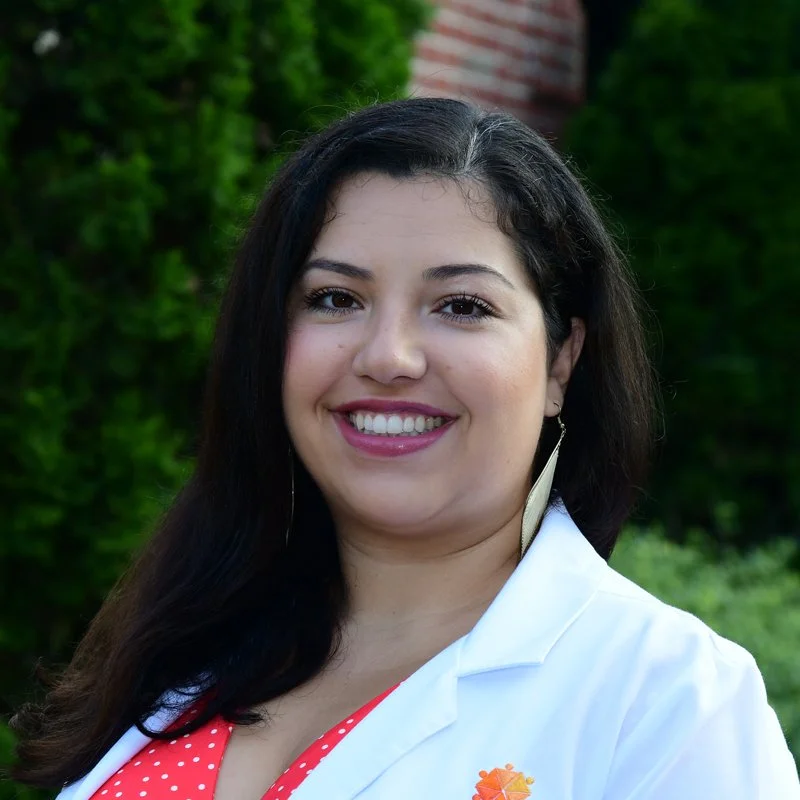 A woman with long dark hair smiling outdoors, wearing a white blazer over an orange polka dot top, with greenery in the background.