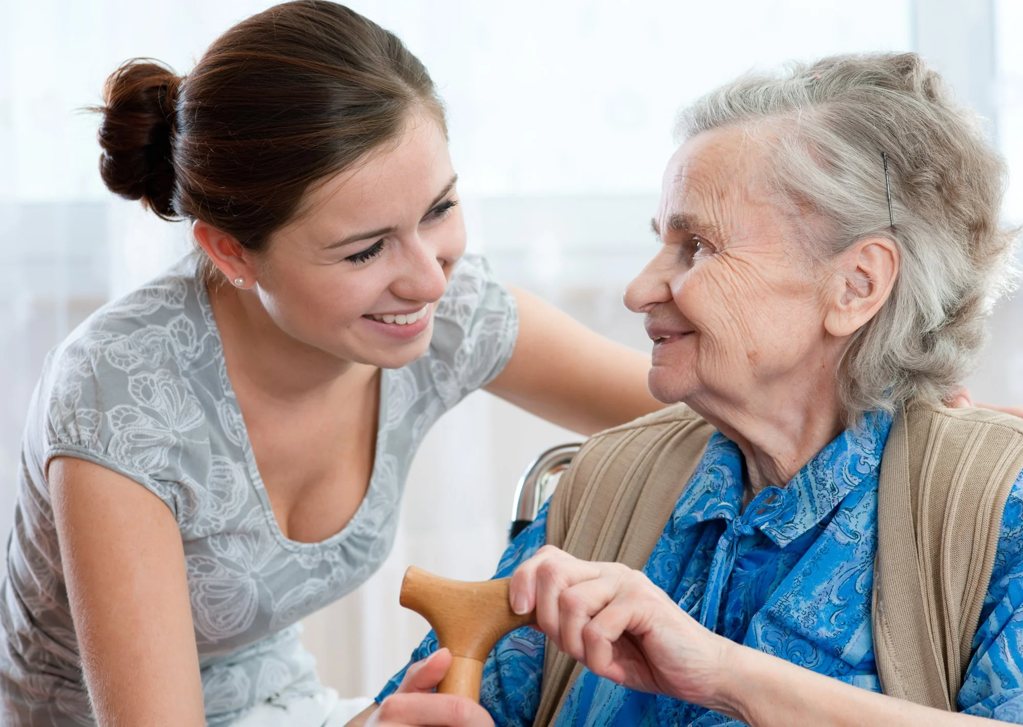 A young woman smiling and leaning toward an elderly woman in a wheelchair, holding a wooden cane, in a bright room.