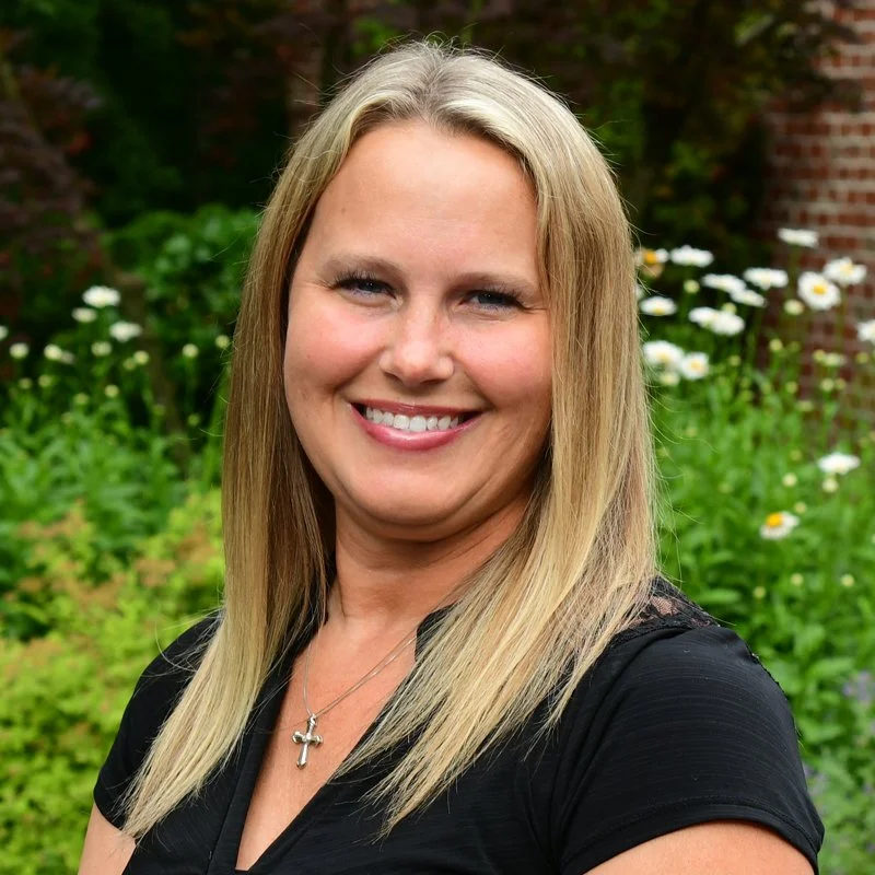 A woman with long blonde hair smiling outdoors, standing in front of green foliage and white flowers.