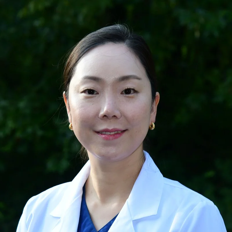 Portrait of a woman with dark hair in a ponytail, wearing a white lab coat and gold earrings, smiling outdoors with green foliage in the background.