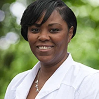 Portrait of a smiling woman with short black hair, wearing a white blazer, outdoors with green foliage in the background.