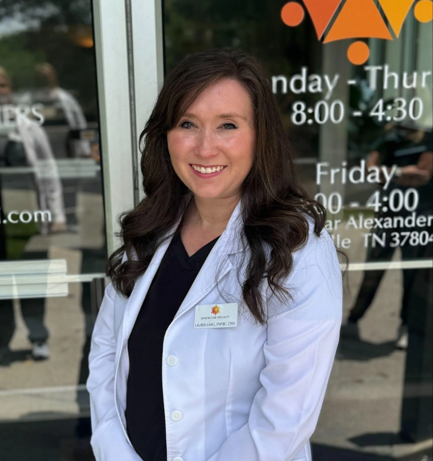 A woman with long brown wavy hair smiling, wearing a white medical coat with a name tag, standing in front of a glass door with business hours and contact info on it.